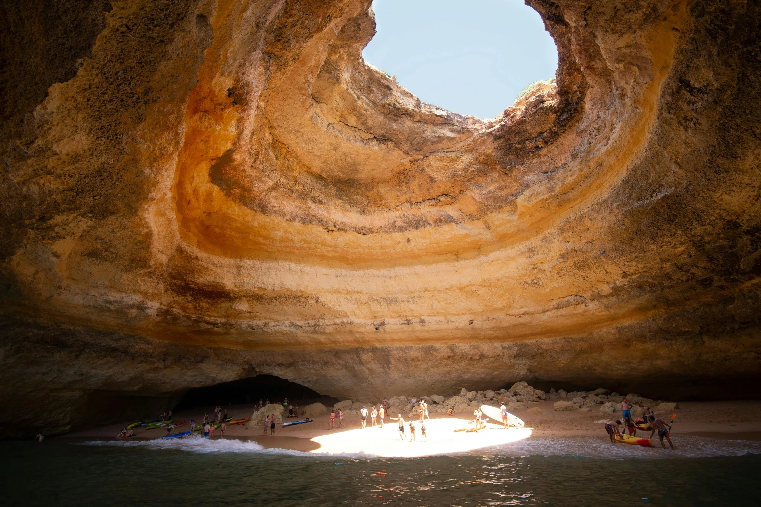 A large sea cave with a sandy floor and a circular opening at the top, sunlight illuminating part of the sandy area inside, with people and kayaks on the sand near the water.