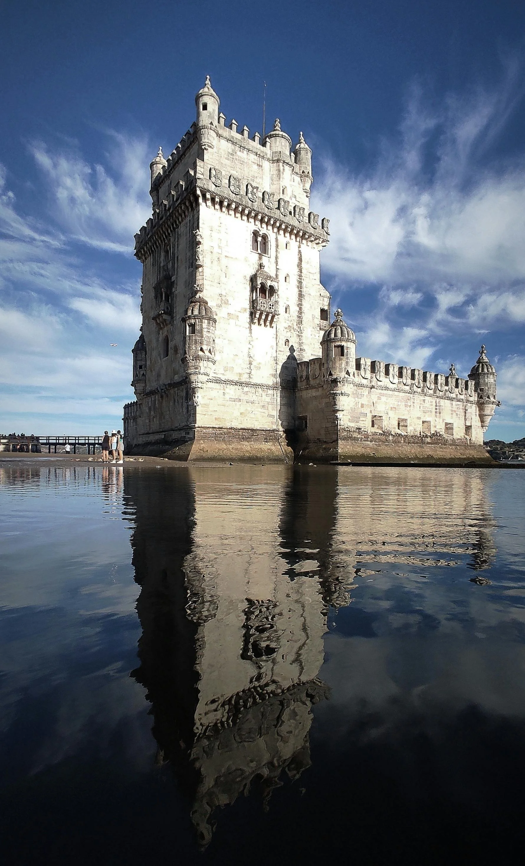 A historic castle with turrets and battlements, surrounded by water reflecting the structure and a partly cloudy blue sky.