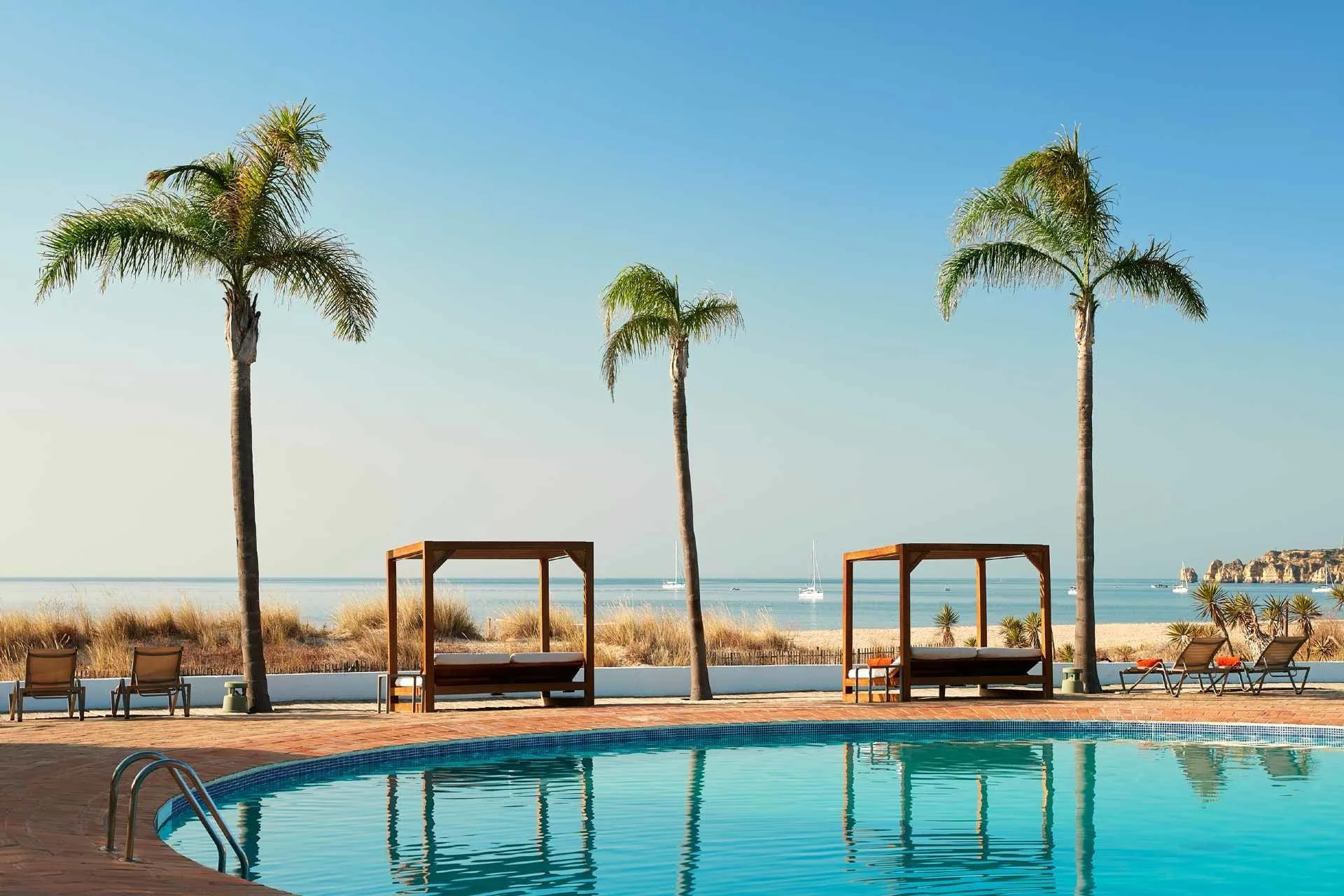 Swimming pool with lounge chairs, cabanas, palm trees, and ocean view with boats in the background.