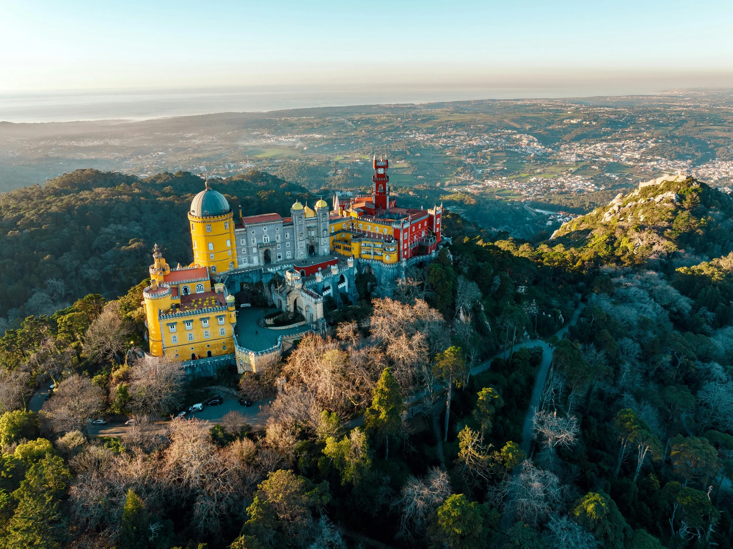 Colorful Pena Palace on a hillside overlooking a forest with a city and ocean in the background.