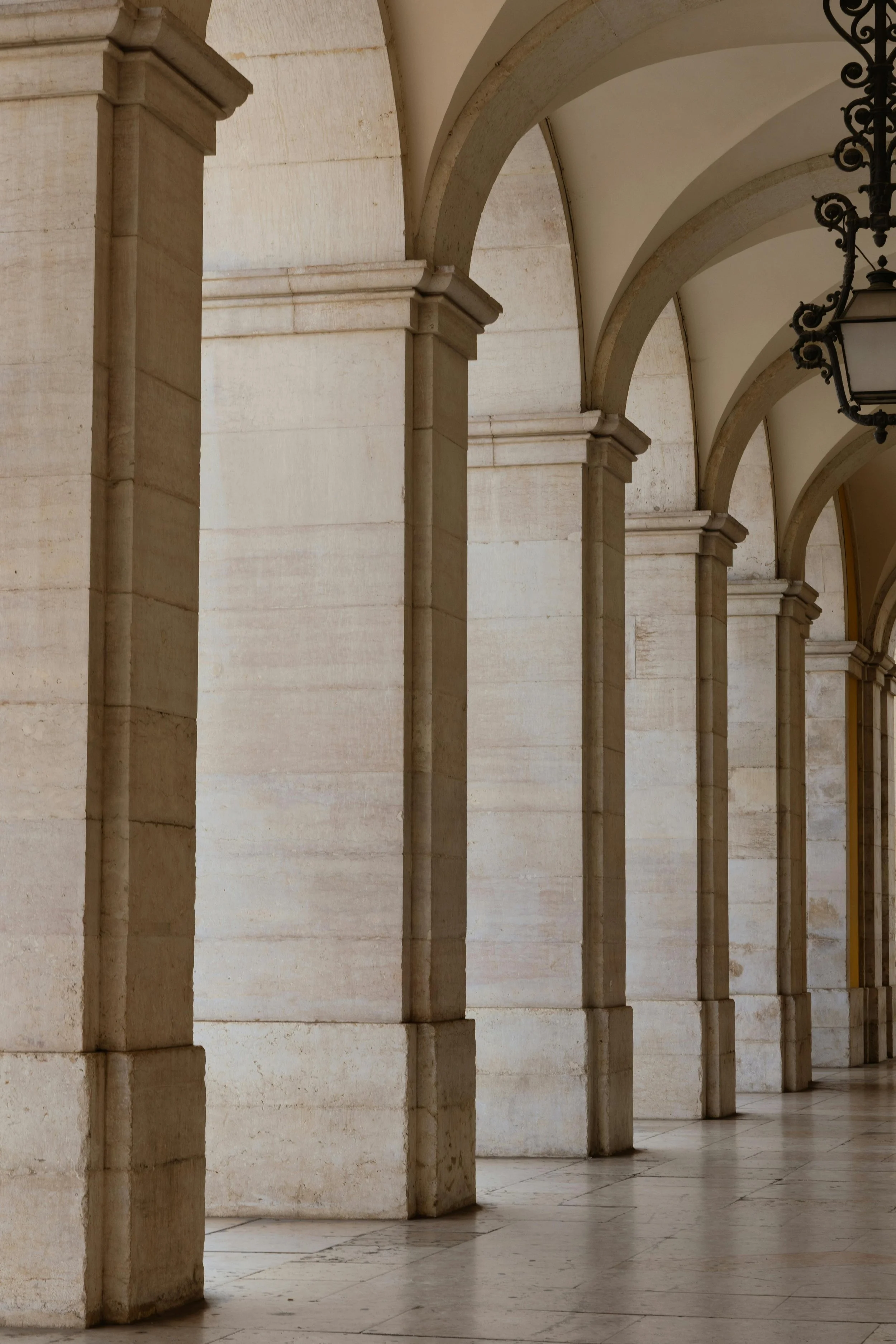 A covered walkway with stone arches and columns, hanging lanterns, and a tiled floor.