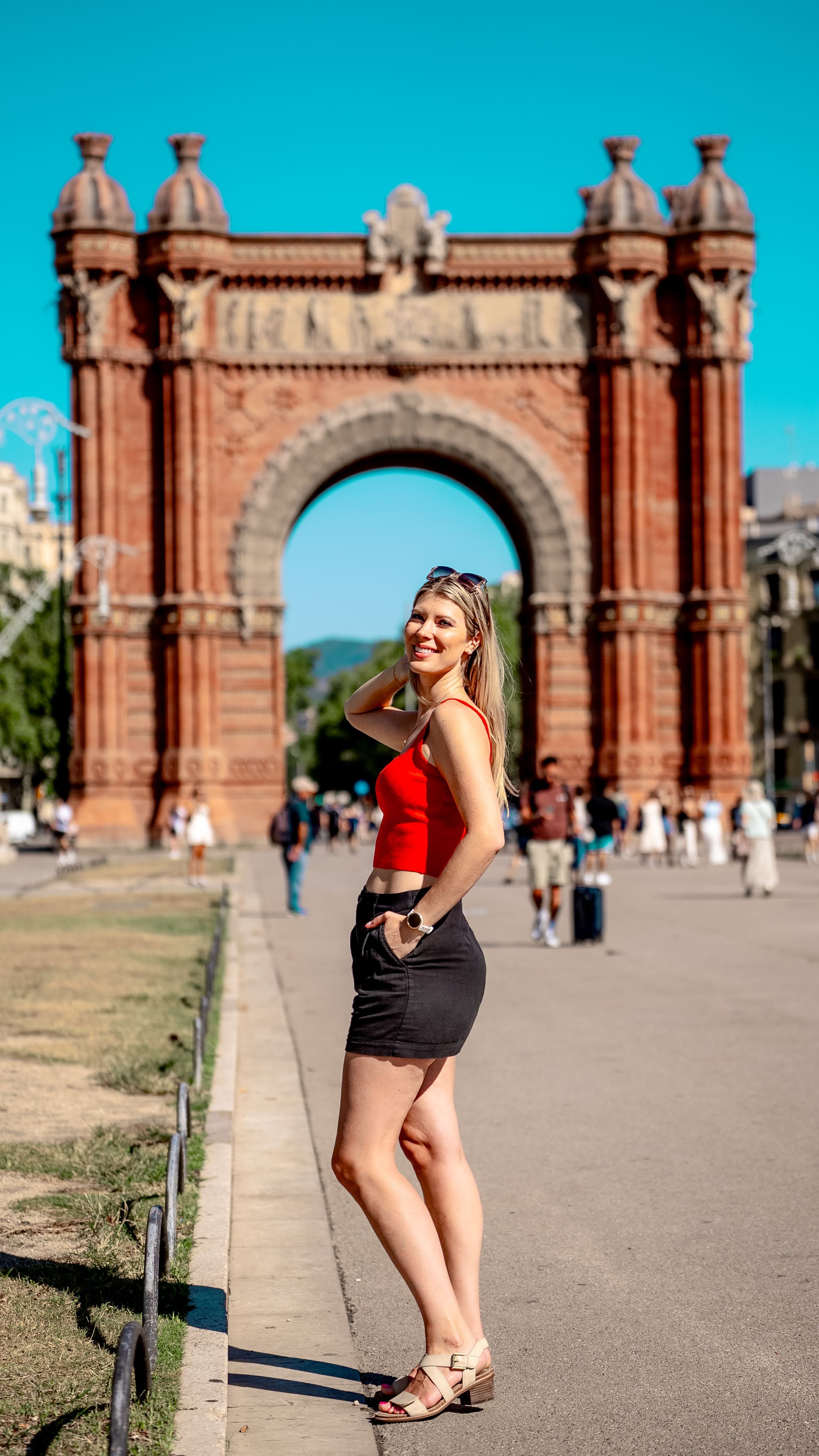 A woman in a red crop top and black skirt standing near the Arc de Triomphe in a busy city square on a sunny day.