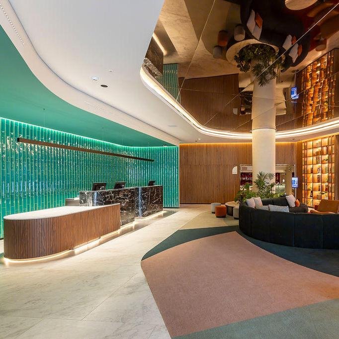 Hotel Mundial Lisbon lobby featuring a curved reception desk, green accent wall with illuminated glass tiles, a white marble reception counter, and a sitting area with a curved sofa, armchairs, and a large branch and potted plants.