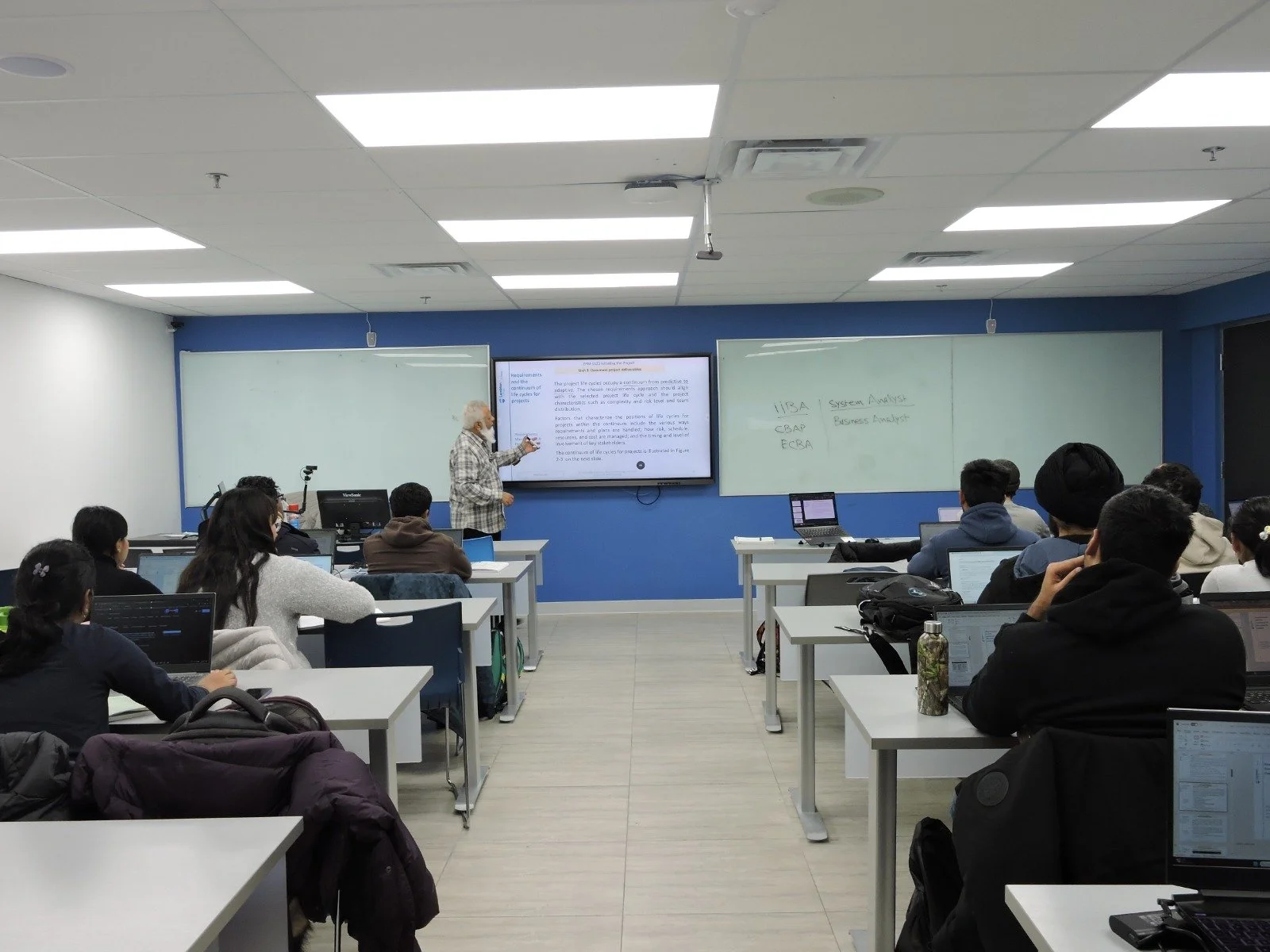 A classroom with students seated at desks listening to a teacher who is standing at the front, pointing at a large screen displaying a presentation, with whiteboards on either side.