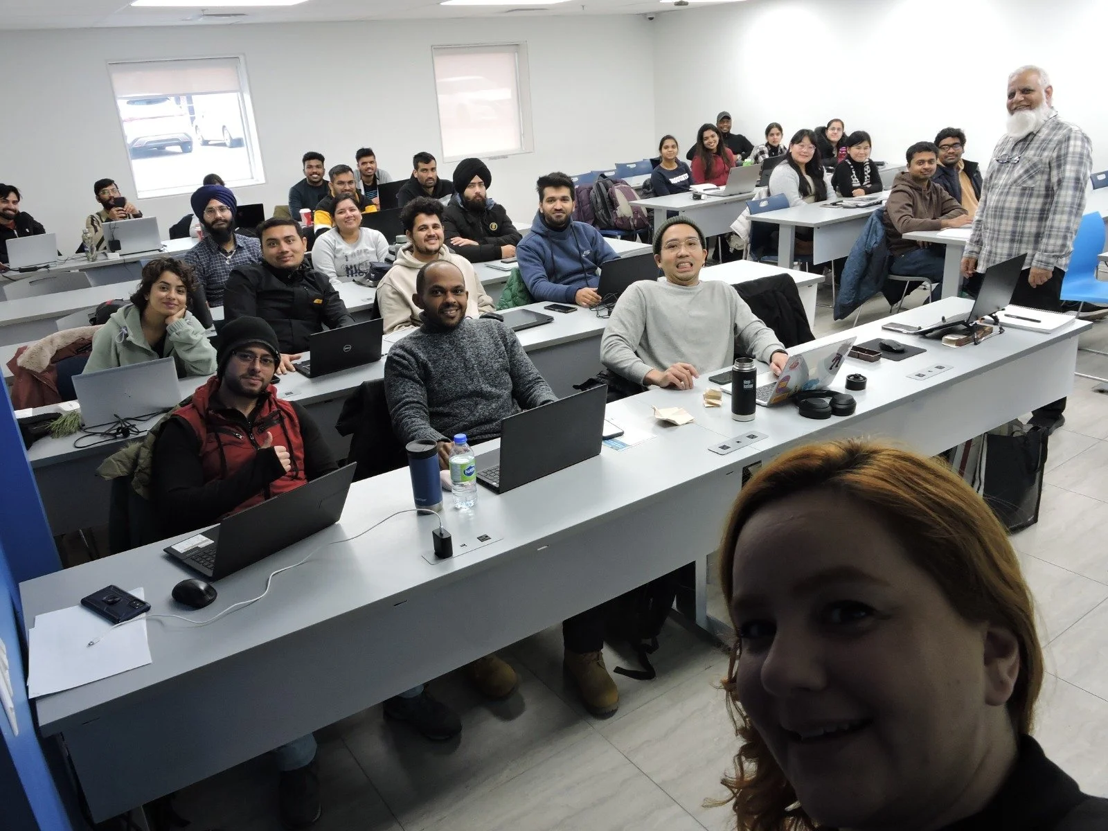 Classroom filled with students and a teacher, with laptops, notebooks, and water bottles on the desks, smiling and looking at the camera.