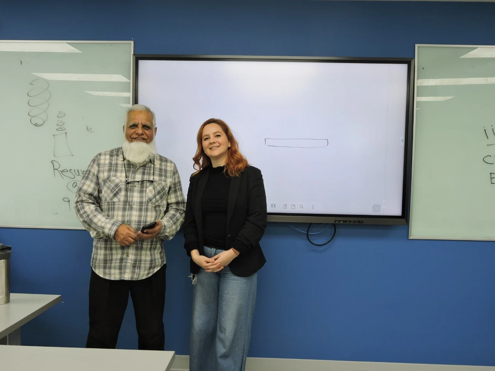 A man with white hair and a white beard wearing a plaid shirt and glasses hanging around his neck, and a woman with wavy red hair wearing a black blazer and jeans, standing together in a classroom in front of a digital whiteboard and whiteboards with
