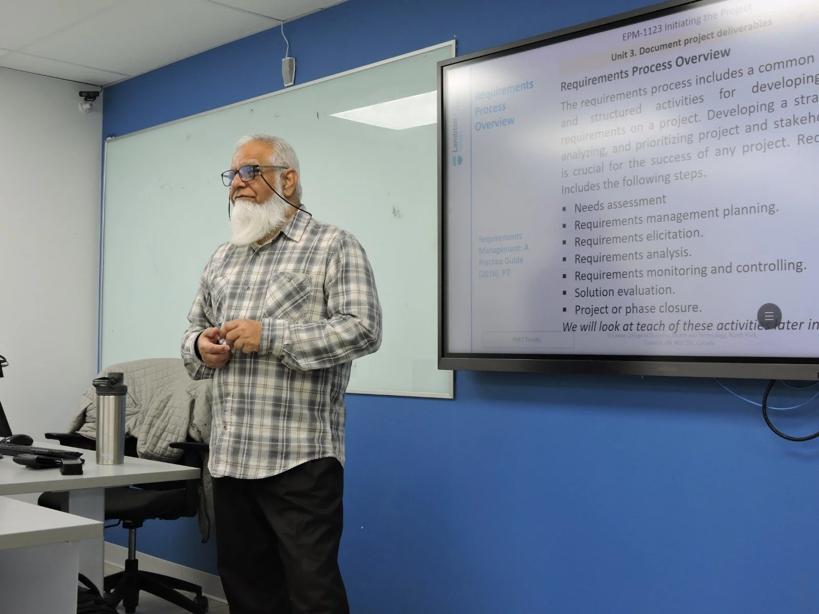An elderly man with gray hair and beard wearing glasses, a plaid shirt, and black pants, standing in front of a whiteboard and a large screen displaying a presentation titled 'Requirements Process Overview' in a classroom or conference room.