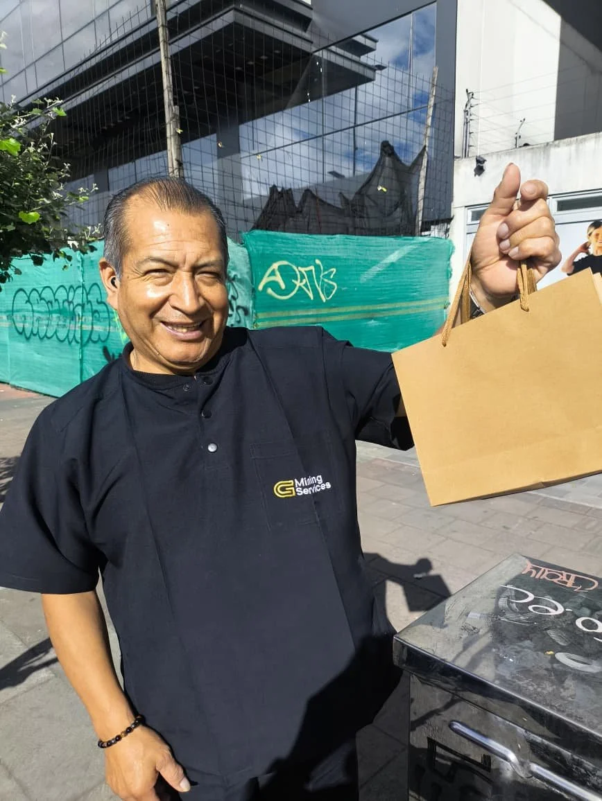 Un hombre sonriendo sosteniendo una bolsa de papel, lleva puesto un uniforme negro con el logo de servicios mineros en el pecho, está en la calle frente a un edificio y una estructura de cartón o plástico verde.