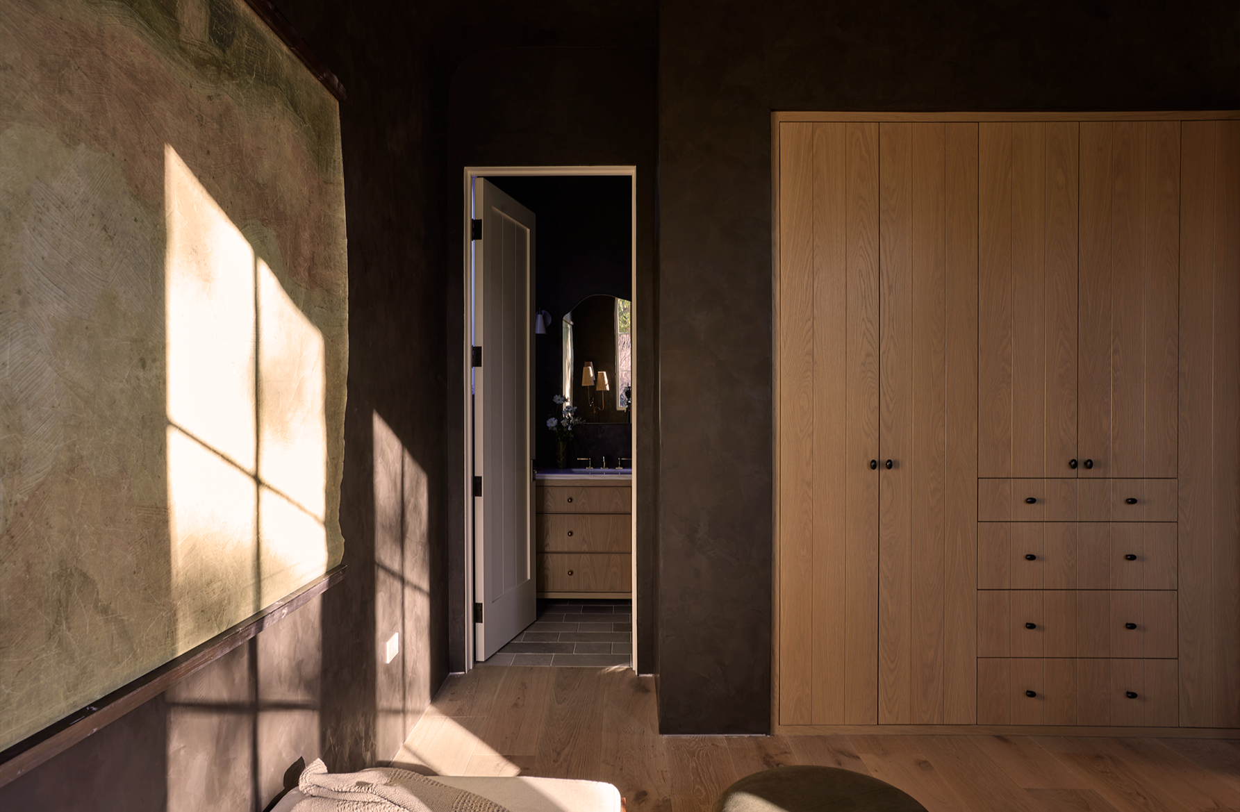 Sunlight streaming through a window creates a shadow pattern on a textured wall in a room. A doorway opens to a bathroom with a vanity and mirror. There is wood cabinetry on the right side of the room, and part of a couch and an ottoman are visible in the foreground.