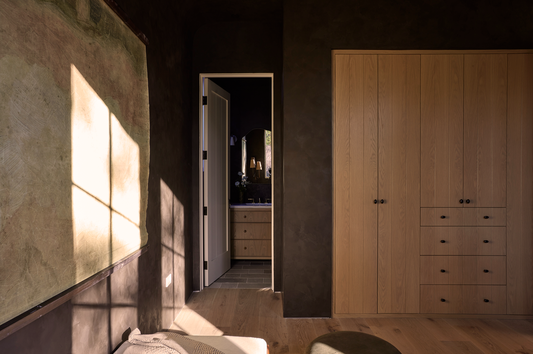 4338 Rhodes Ave, Studio City. Sunlight casting a shadow of a window onto a textured wall in a cozy bedroom, with a door leading to a bathroom, and wooden furnishings.
