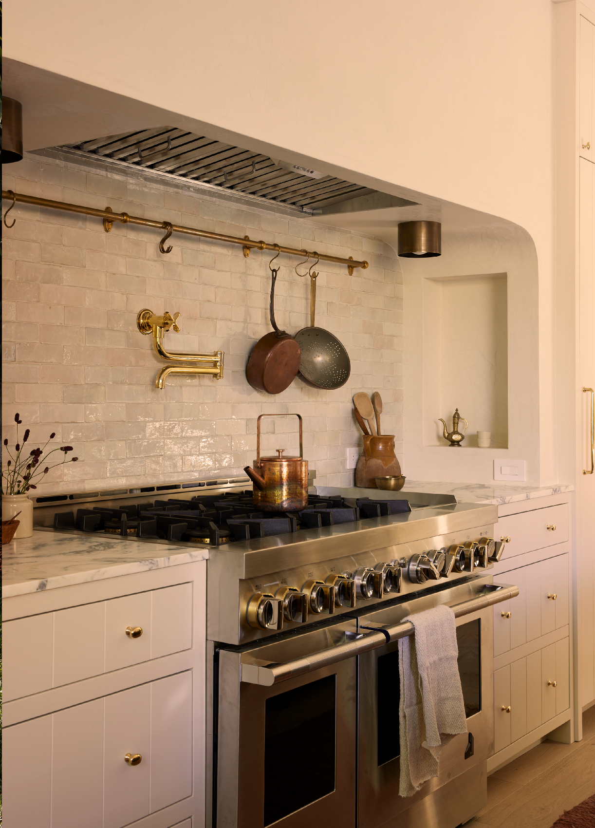 Kitchen with a stainless steel gas stove, cream cabinetry with gold knobs, white tile backsplash, hanging copper and metal pots, a brass faucet, marble countertop, and a towel hanging from the stove handle.