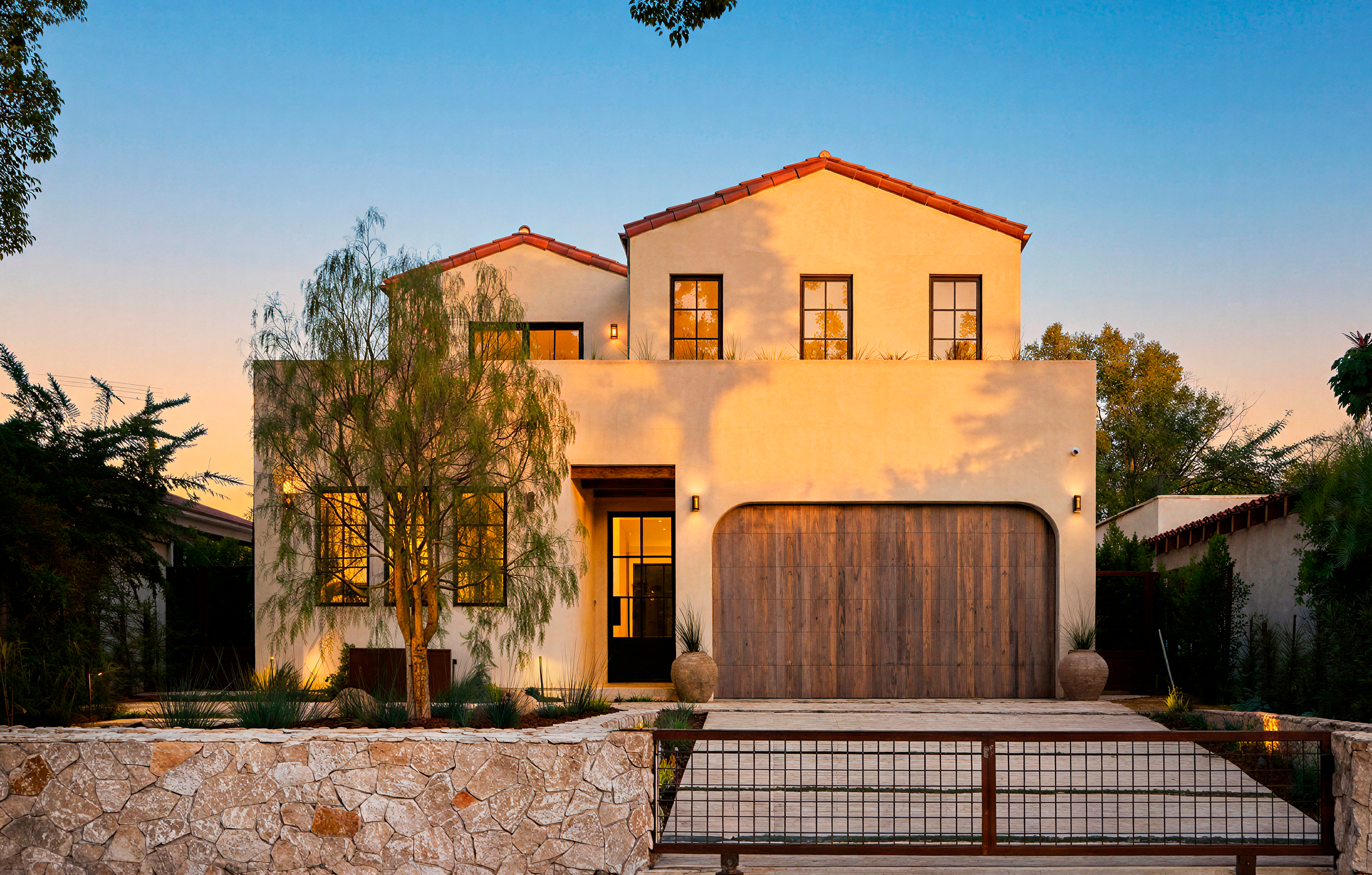 4338 Rhodes Ave, Studio City. Modern two-story house with white exterior, large wooden garage door, and illuminated windows during sunset, surrounded by trees and landscaped yard.