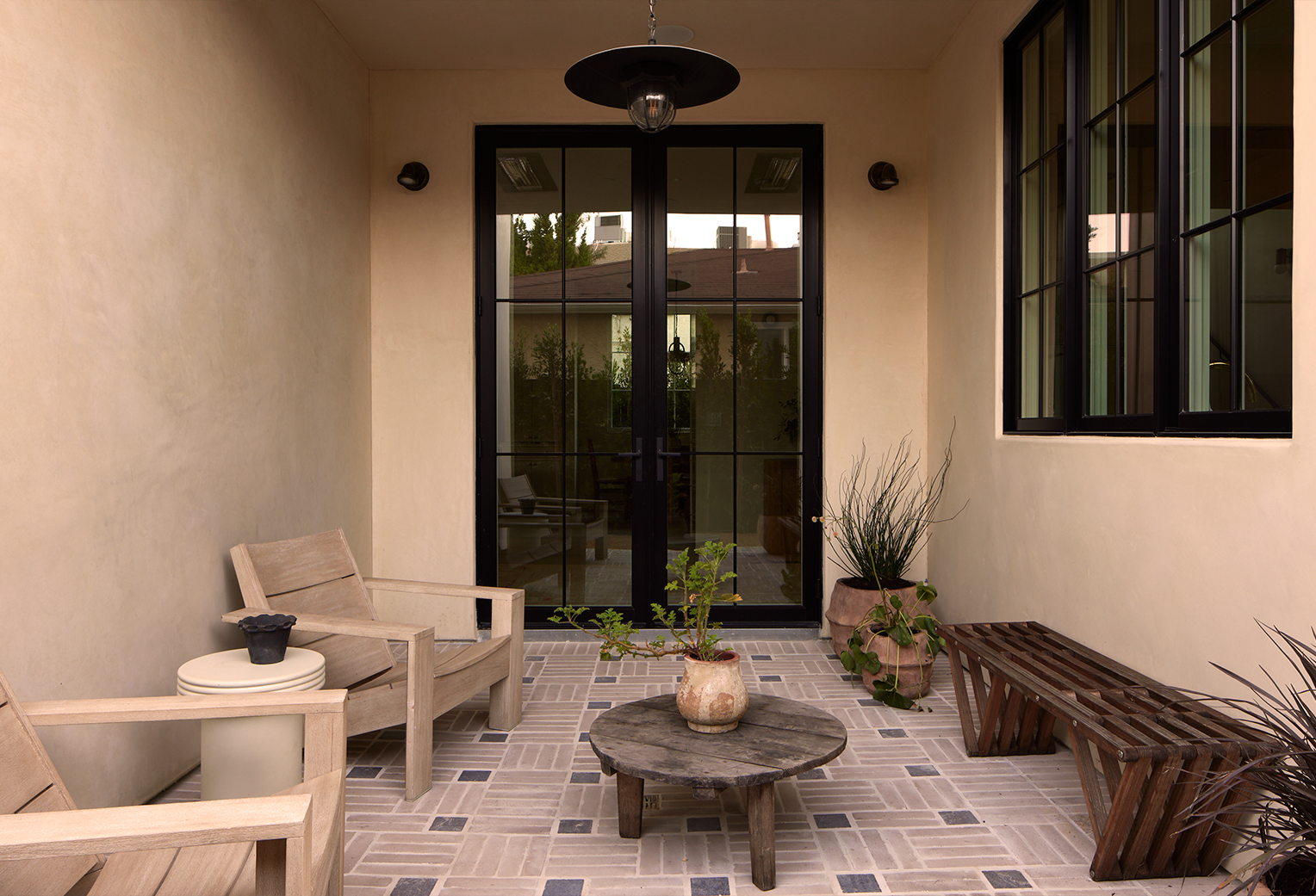 A small outdoor patio area with beige walls and black-framed glass doors leading inside. There are two beige armchairs with a small round side table between them on the left. In the center, there is a low round wooden coffee table with a potted plant. To the right, a wooden bench and two potted plants are against the wall. The floor is tiled with beige and dark tiles in a geometric pattern.
