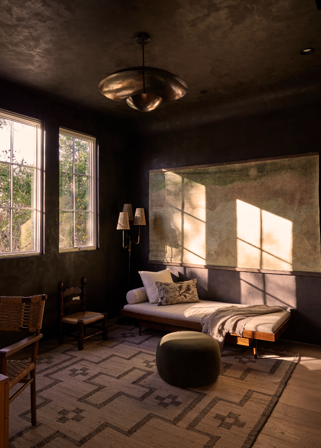 4338 Rhodes Ave, Studio City. bedroom with dark walls and hardwood floors, featuring a bed with pillows and a blanket, two windows letting in natural light, a patterned rug, a small round ottoman, a wall-mounted map, and a floor lamp with two shades.