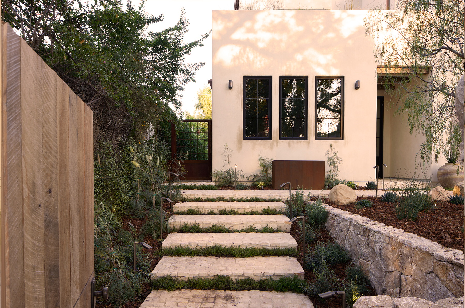 Contemporary house with white stucco exterior, black-framed windows, and wooden landscaping elements. Stone steps lead to the entrance, flanked by plants and short grass. A large decorative stone and potted plants are on the right side. Overhanging trees cast shadows on the house façade.