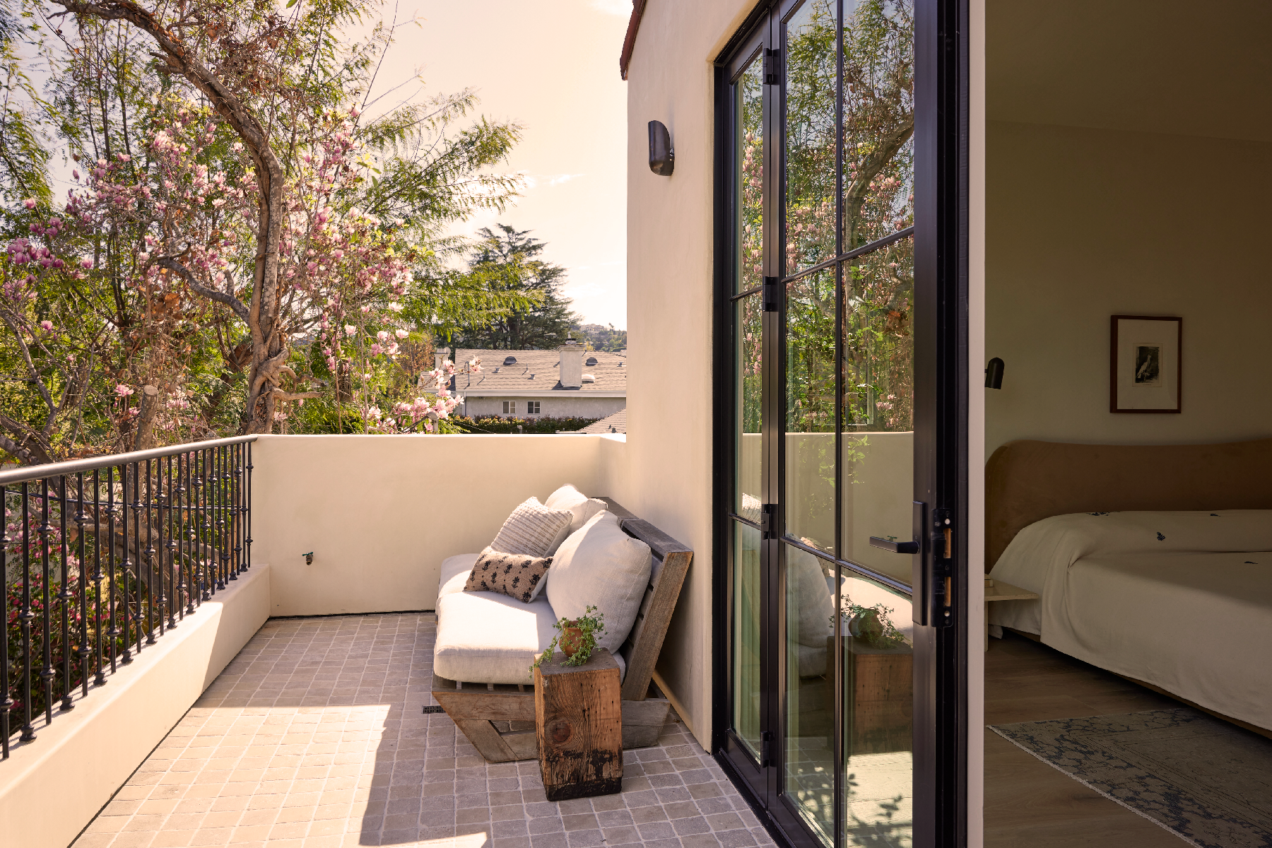 4338 Rhodes Ave, Studio City. View of a cozy balcony with a white couch and decorative pillows, overlooking blooming pink trees and neighboring rooftops, adjacent to a bedroom with sliding glass doors.