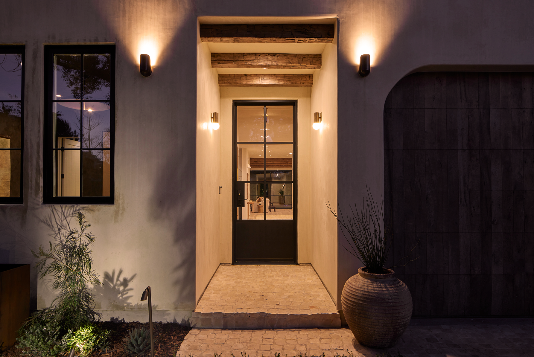 4338 Rhodes Ave, Studio City. Night exterior view of a modern house entrance with a glass door, warm wall lights, potted plants, and a cobblestone pathway.
