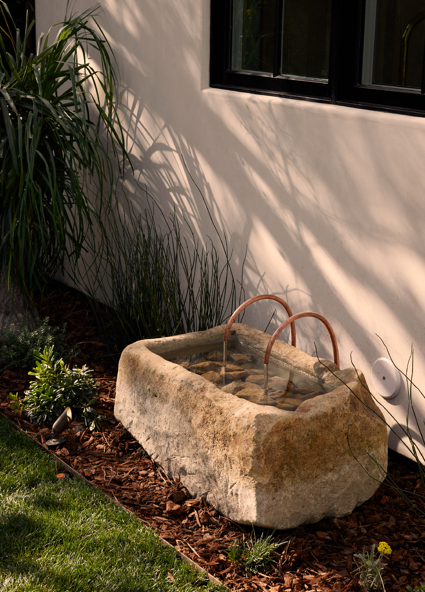 4338 Rhodes Ave, Studio City. A stone water trough with two copper pipes filling it with water, located next to a white exterior wall and surrounded by plants and mulch.