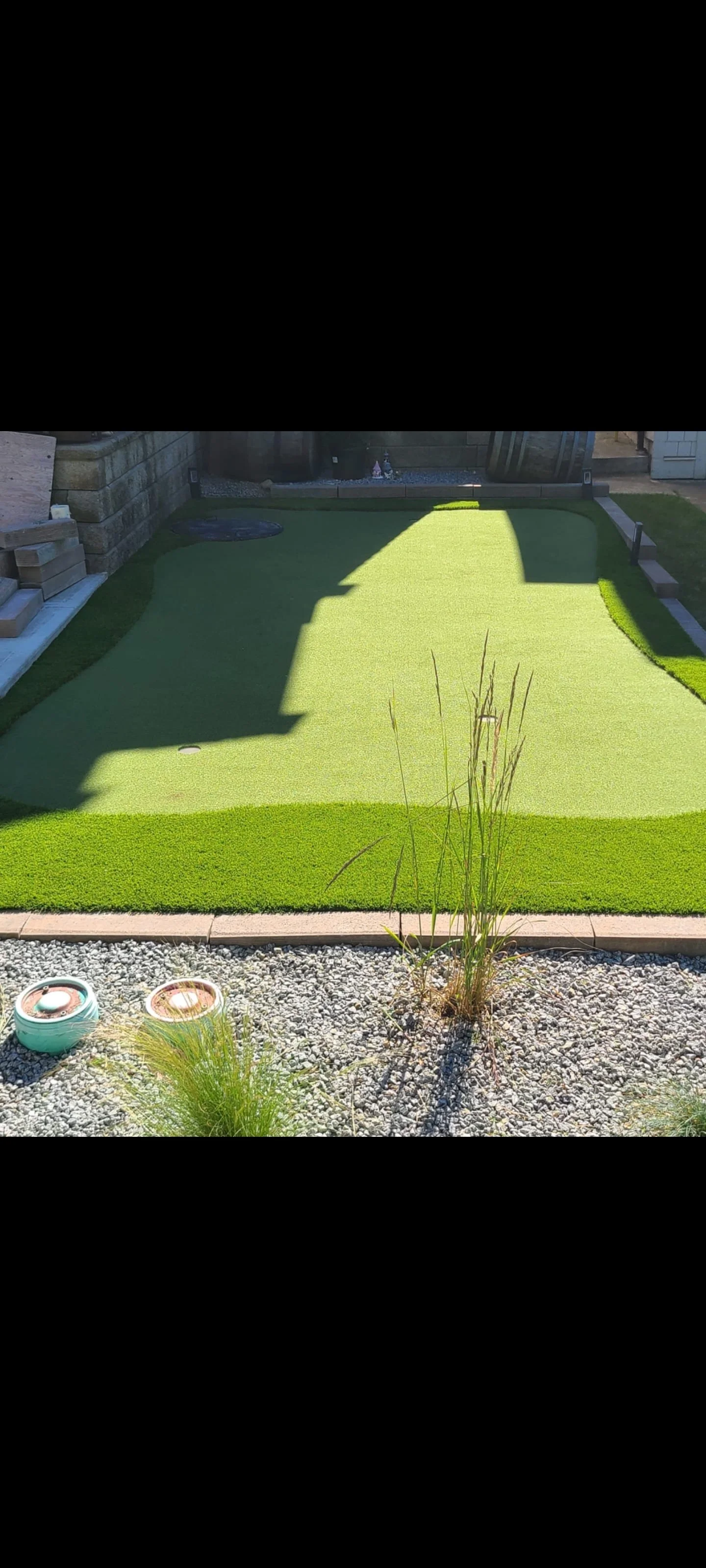 A backyard with a mini golf putting green near langley and surrounding areas, surrounded by gravel and plants, with gardening supplies and barrels in the background.