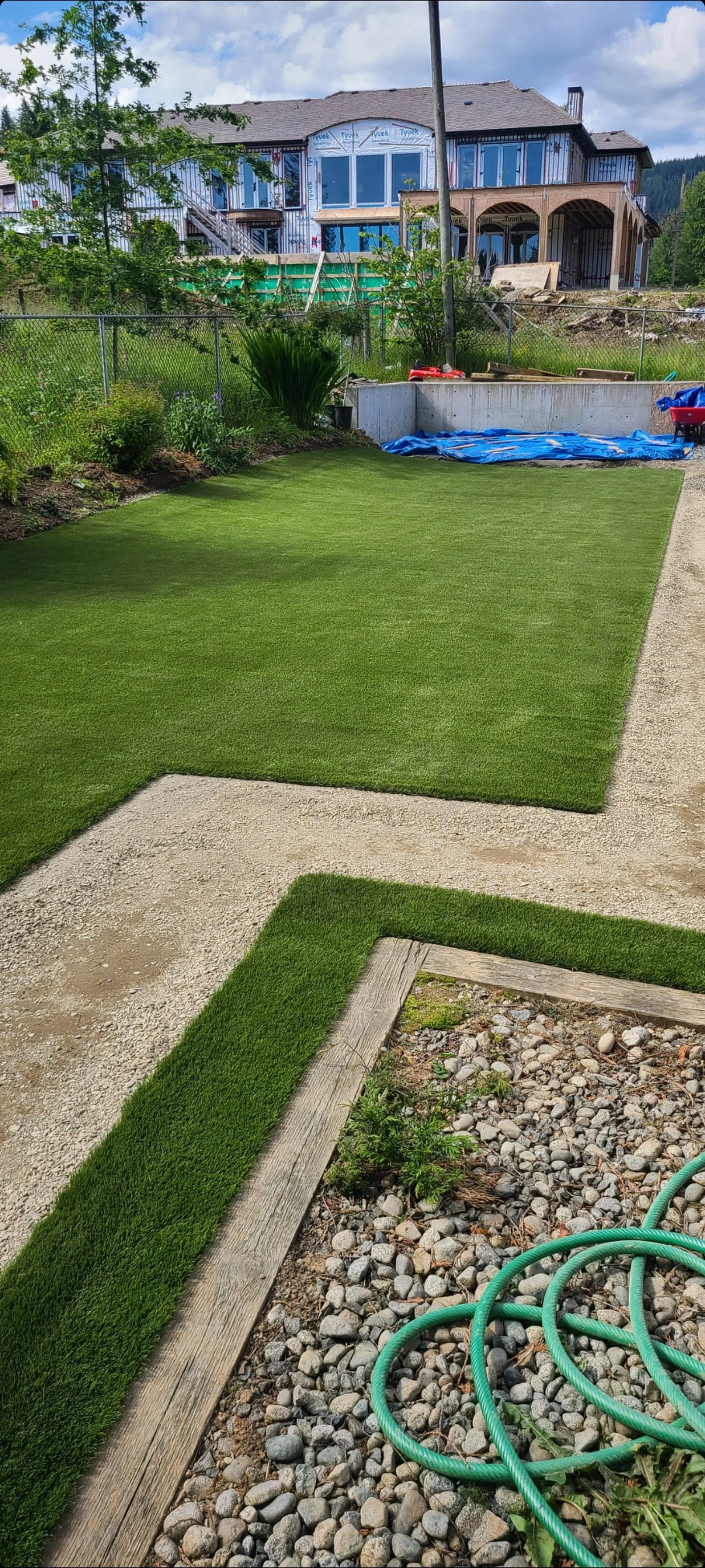 A backyard with a well-maintained lush green lawn, a gravel pathway, and a house under construction in the background. Construction materials and tools are visible.