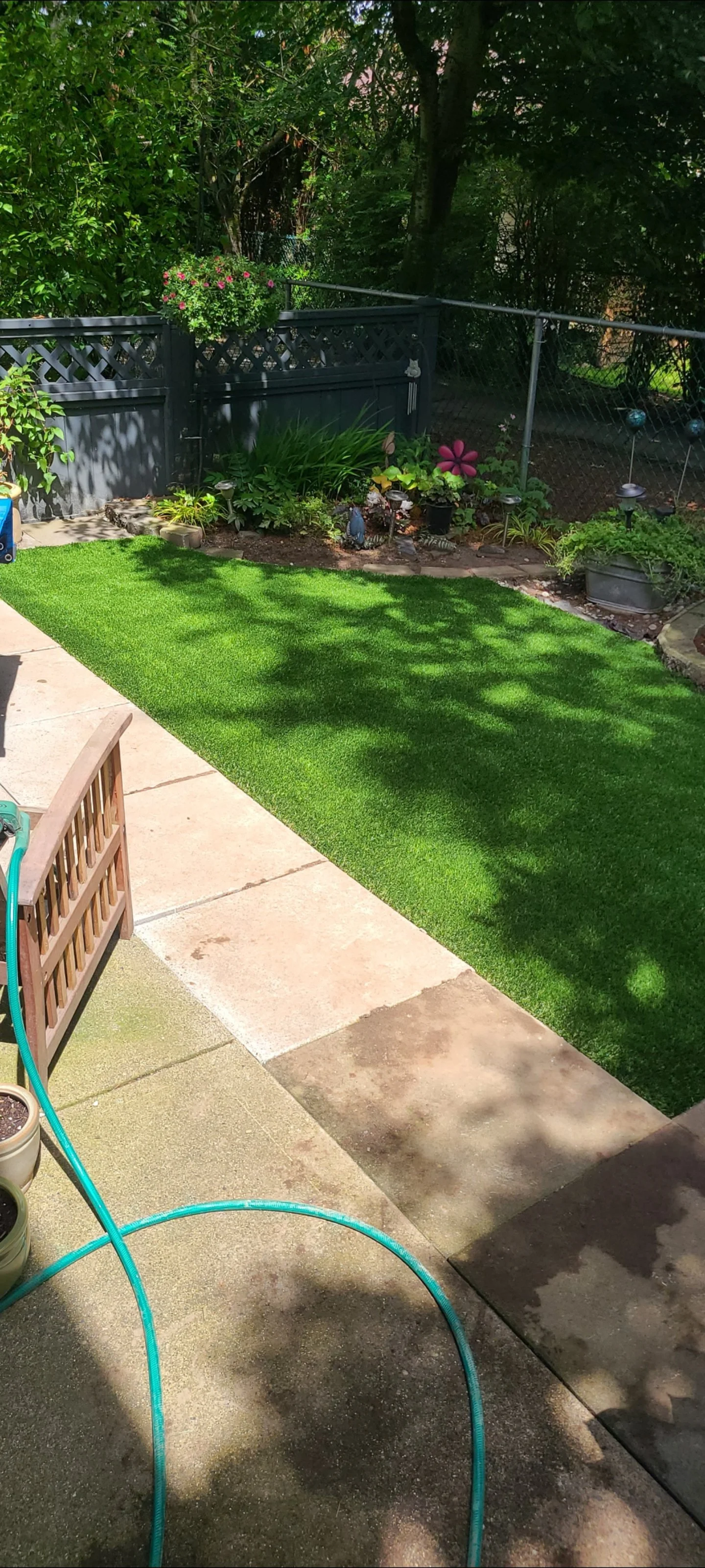 A backyard patio with a garden bed, artificial grass, and a fence. You can see a garden hose on the ground, along with some potted plants and decorative garden ornaments.