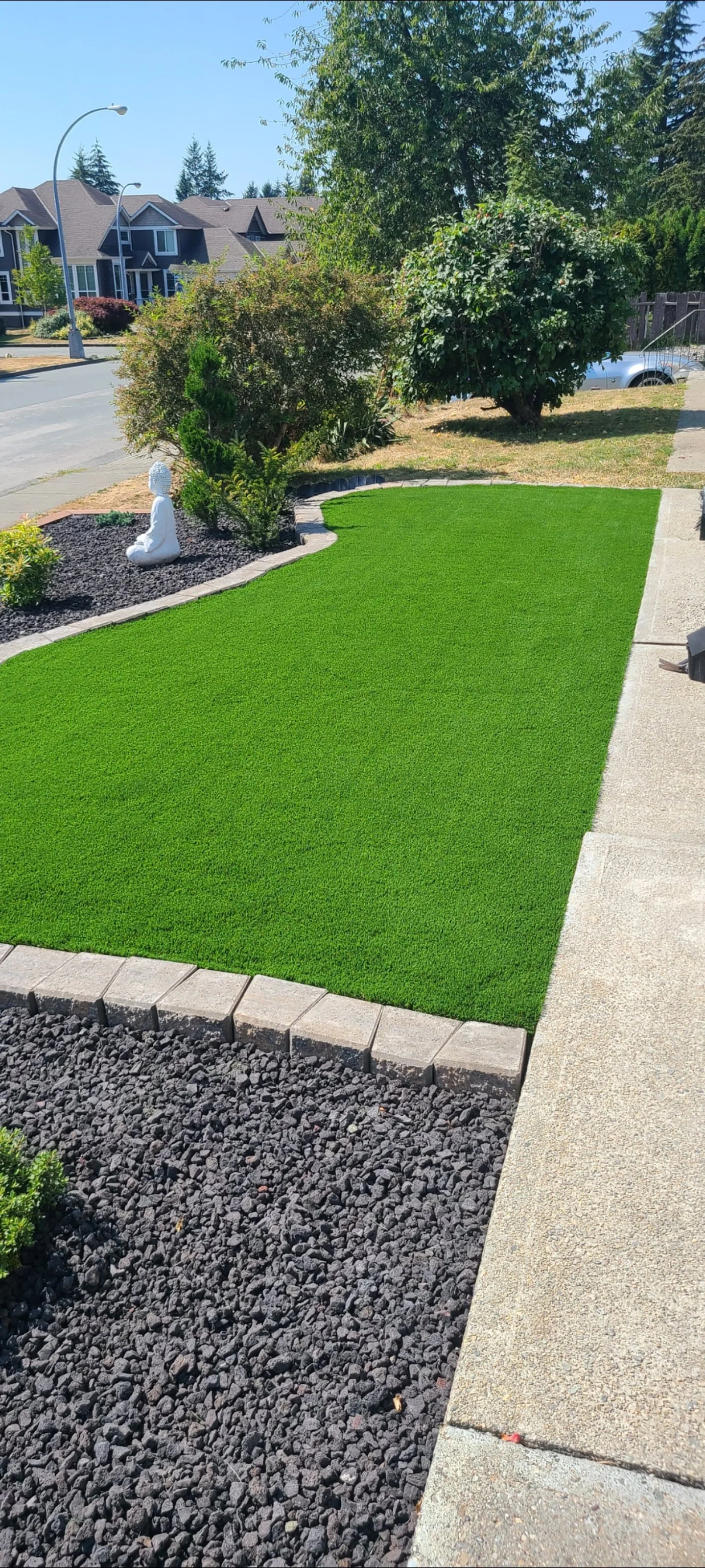 A well-maintained front yard with a small patch of artificial grass, bordered by concrete pavement and black decorative stones, with bushes and trees in the background, and a white Buddha statue on the left side.