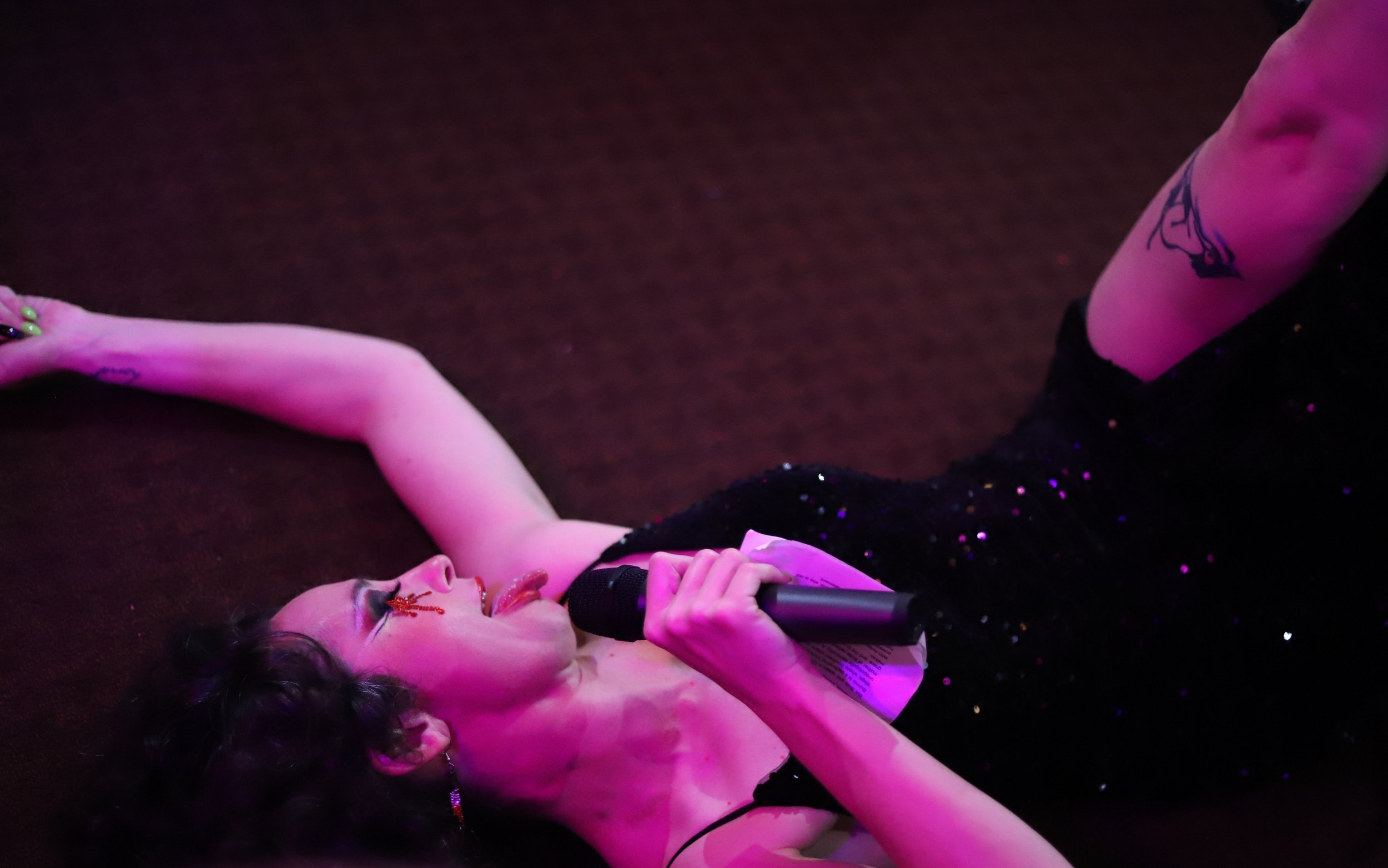 A woman lying on her back on a dark stage, holding a microphone, singing with her eyes closed and her mouth open. She is wearing a black, sparkly dress, with tattoos on her arm and leg, and has dark wavy hair and earrings.