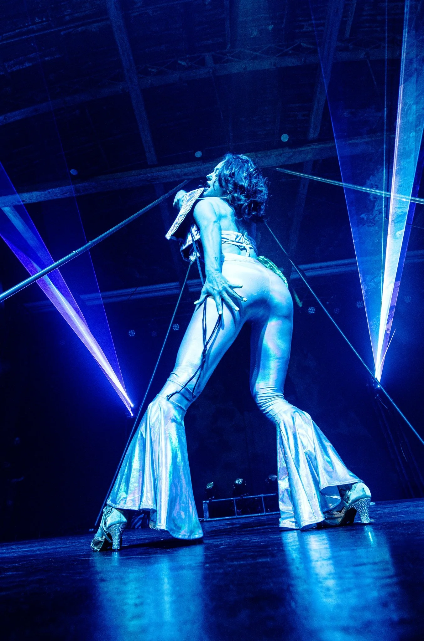 A performer in shiny pants and high heels on stage with dramatic blue lighting and laser effects.