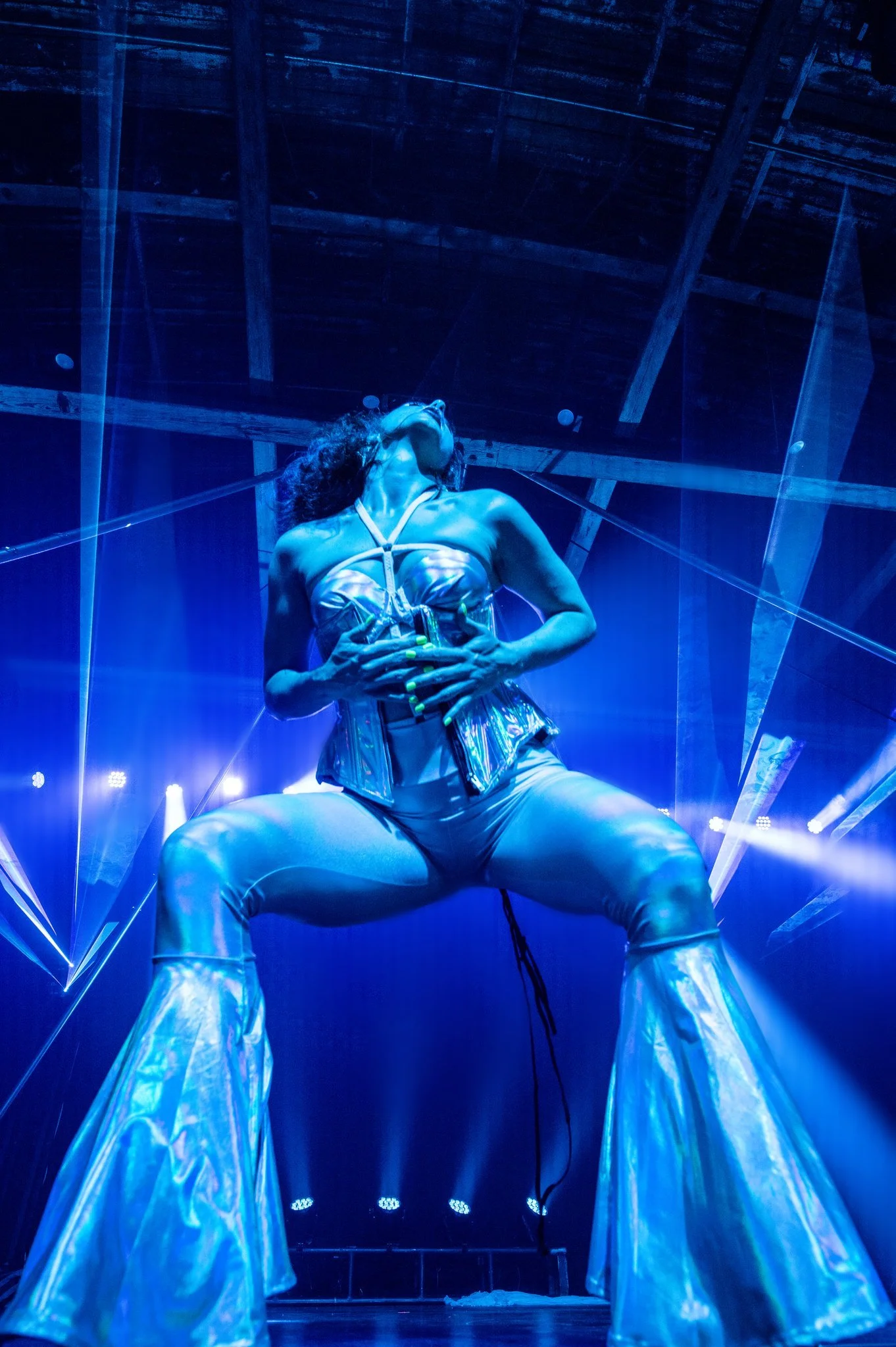 Performer on stage in metallic outfit with blue lighting and laser effects, squatting with legs apart and head tilted back.