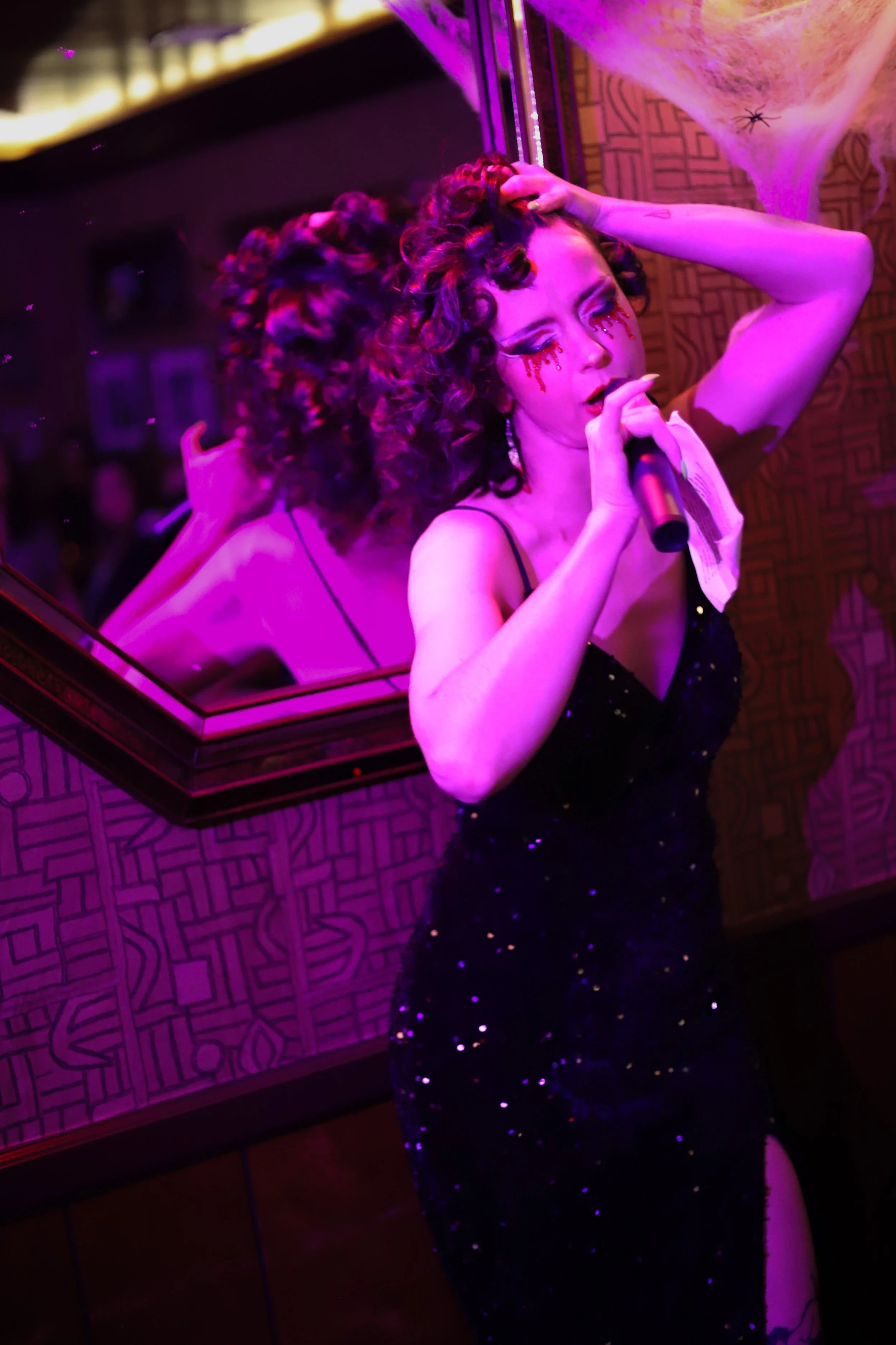 A woman singing into a microphone in a dimly lit room with purple lighting, wearing a black dress with spaghetti straps and sequins, with elaborate curly hair and dramatic makeup, standing in front of a mirror that shows her back and reflection.