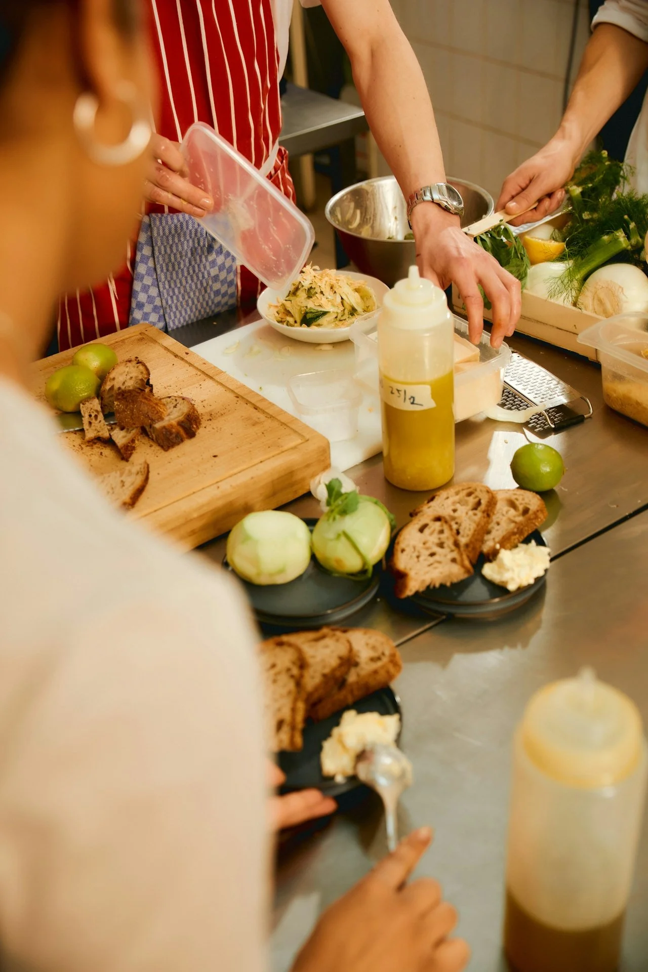 People preparing food in a kitchen, slicing bread, and arranging ingredients on a countertop with various vegetables, bread, and condiments.