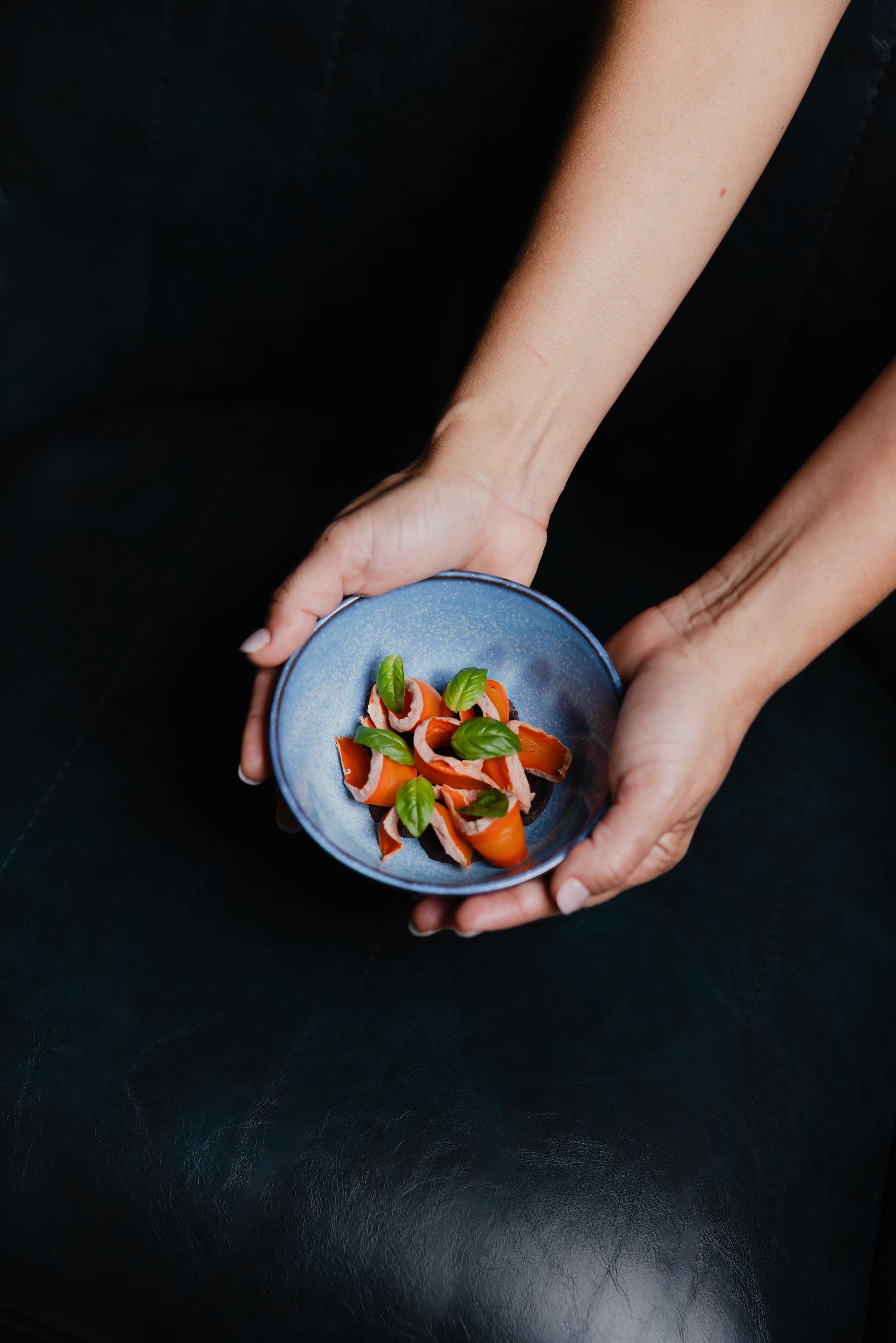 A person holding a small blue bowl with spiral slices of smoked salmon garnished with fresh basil leaves on a dark surface background.