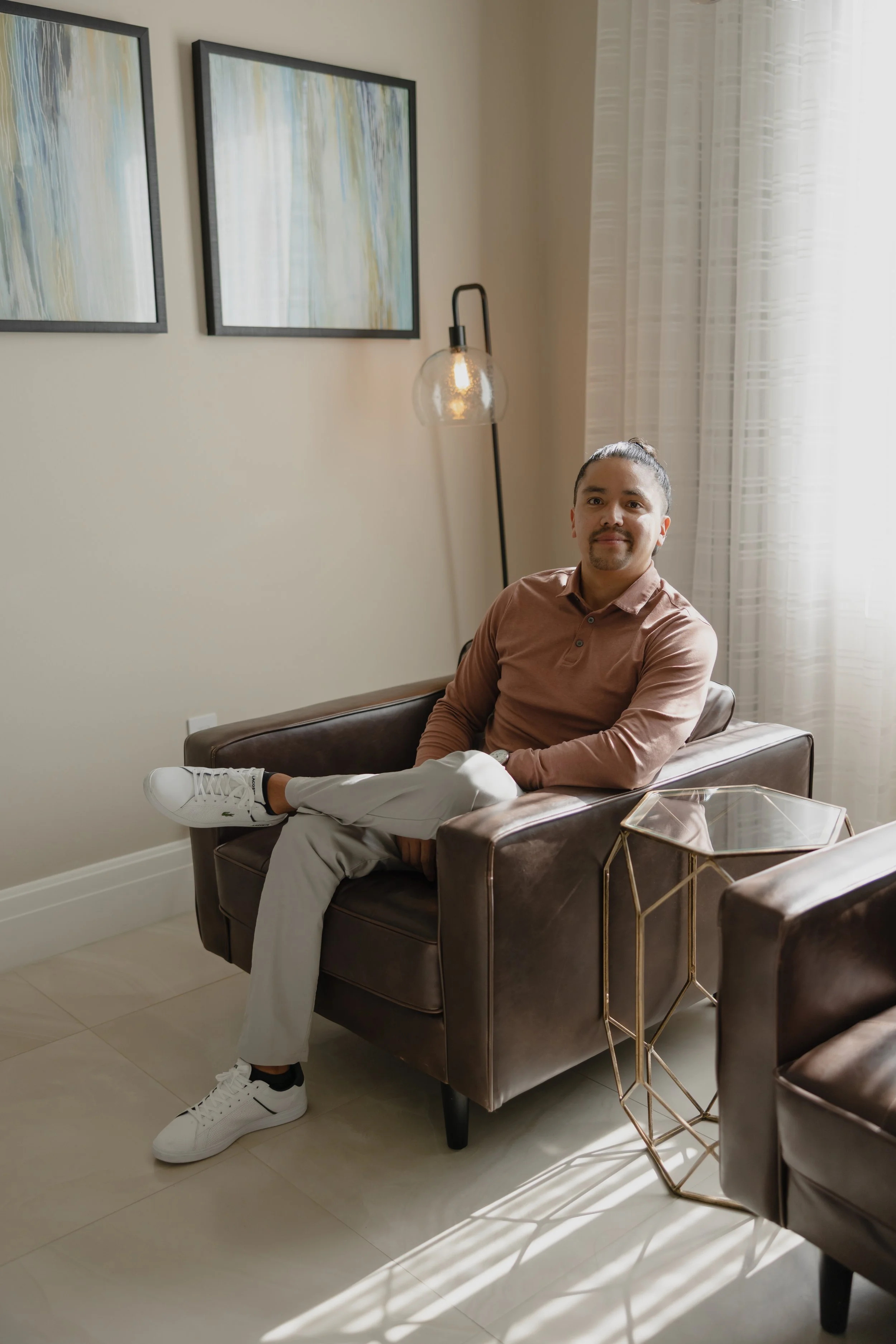 A man with long hair tied back, beard, and mustache is sitting on a brown leather armchair in a modern living room, wearing a light brown shirt, white pants, and white sneakers, with sunlight coming through sheer curtains.