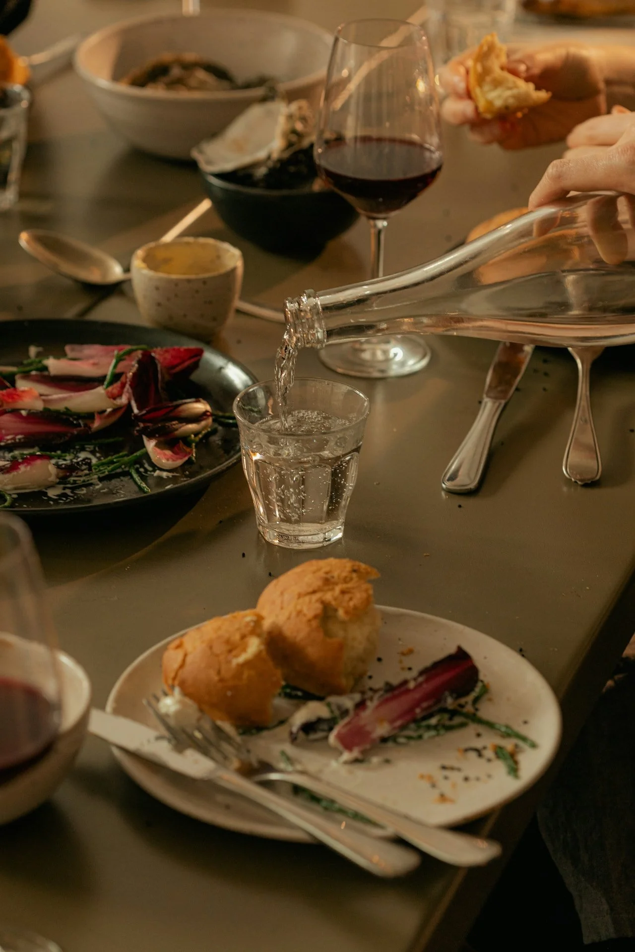 A dinner table with a glass of red wine, a plate with fried chicken and salad, a glass of water being poured from a bottle, and various dishes and utensils on the table.