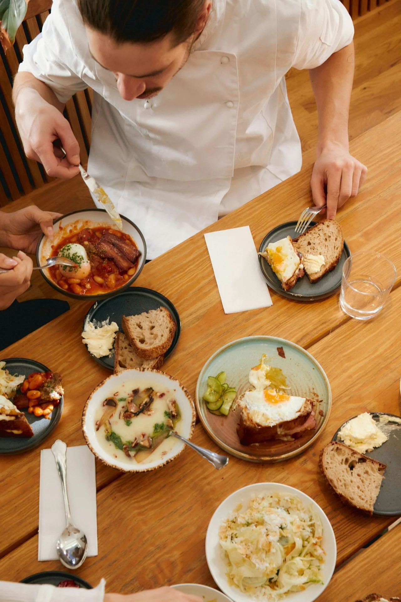 A dining table with various dishes, including bread, eggs, stew, salad, and other side dishes, with a person wearing a white shirt serving food.
