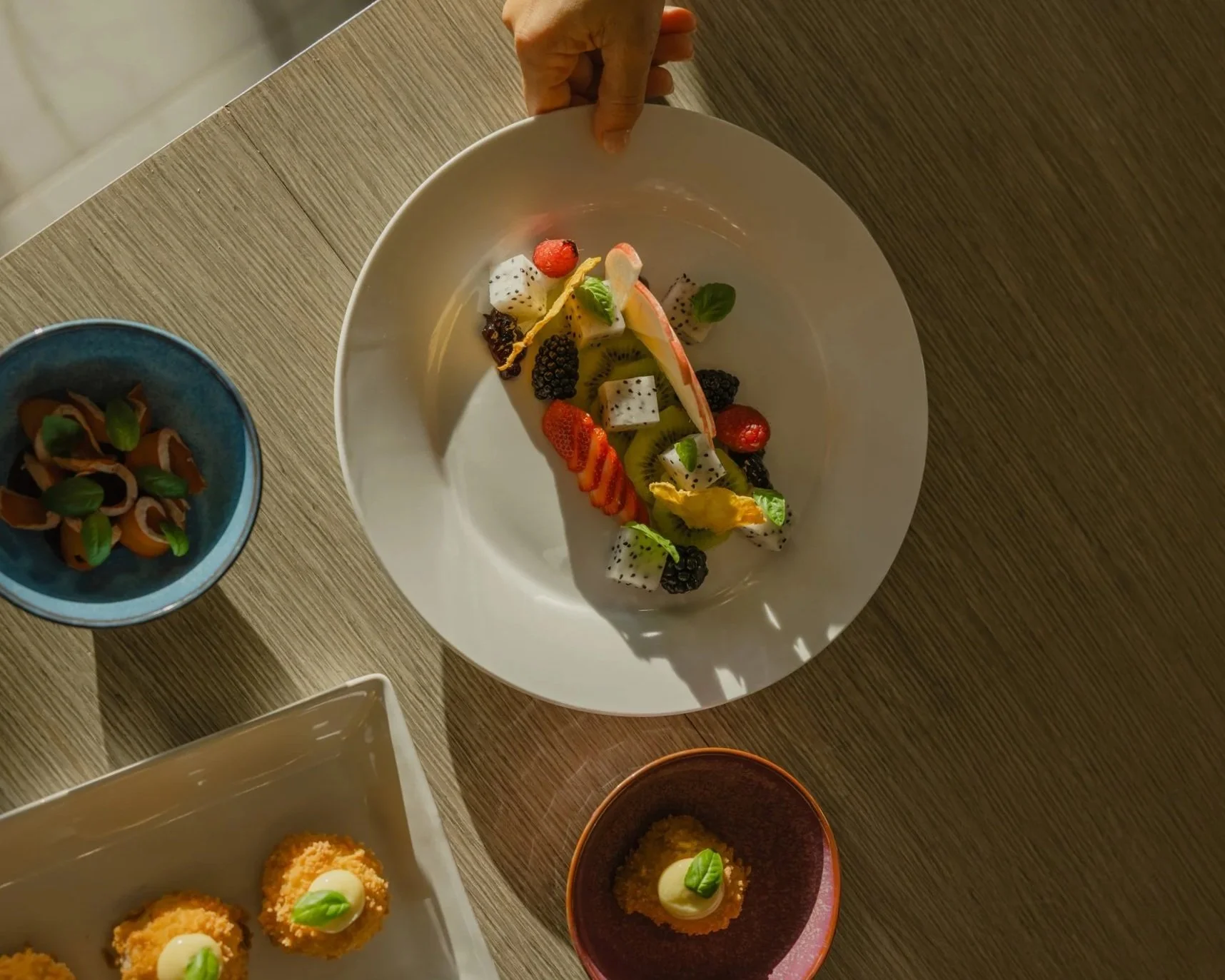 Top-down view of a dessert plate with fruit pieces including strawberries, blackberries, kiwi, dragon fruit, and apple slices, garnished with small mint leaves on a wooden table.