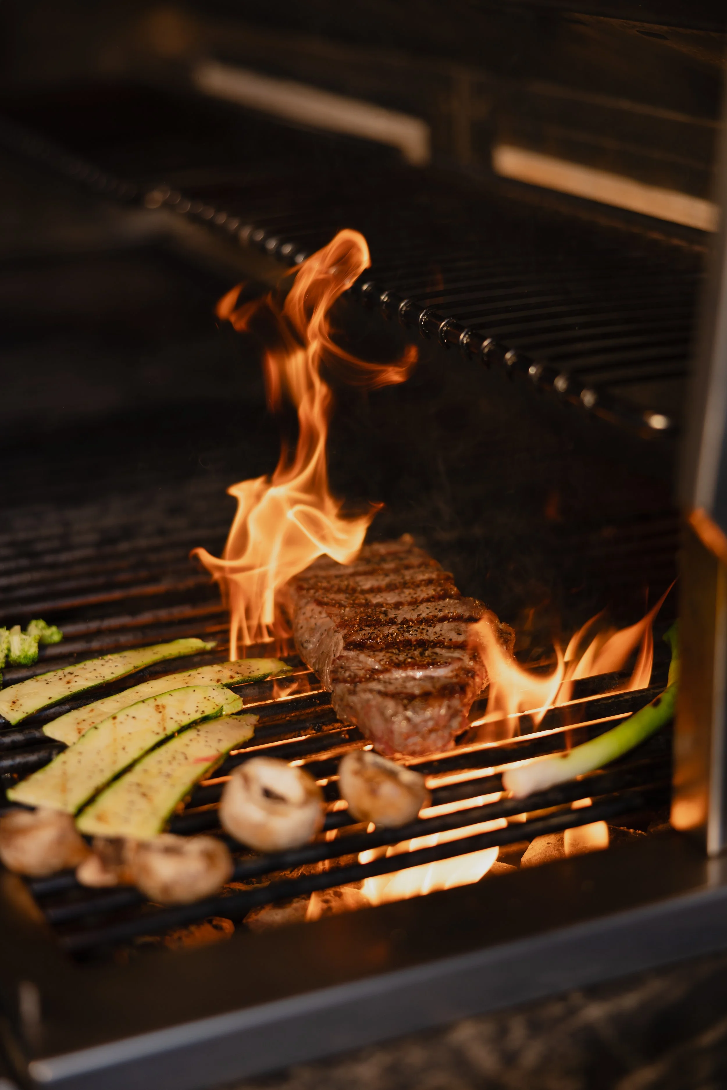 A grill with flames, cooking a steak, zucchini slices, and mushrooms.