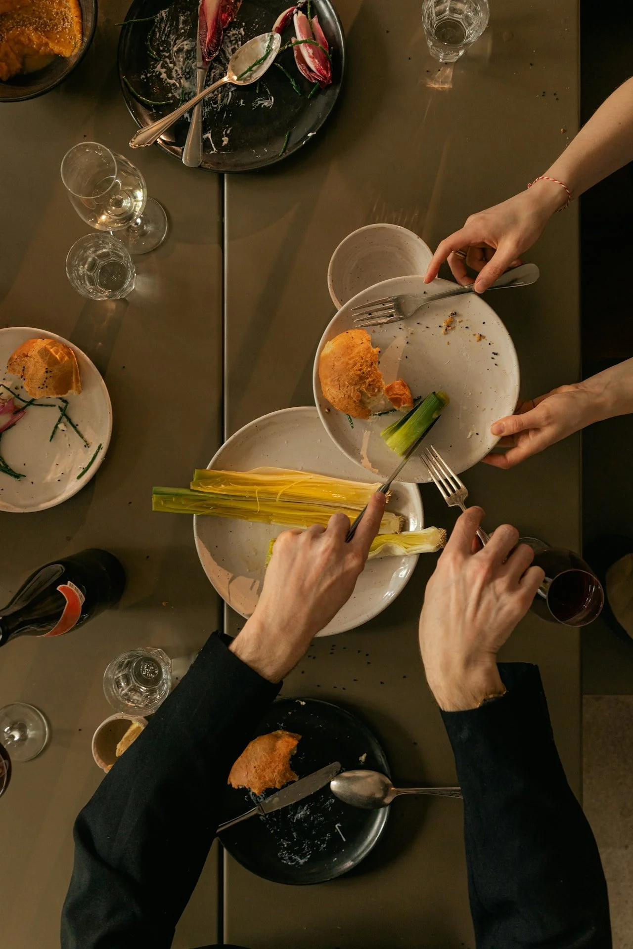 Empty plates with leftover food, wine glasses, and partially eaten bread on a dark dining table, with multiple people reaching for plates and utensils.