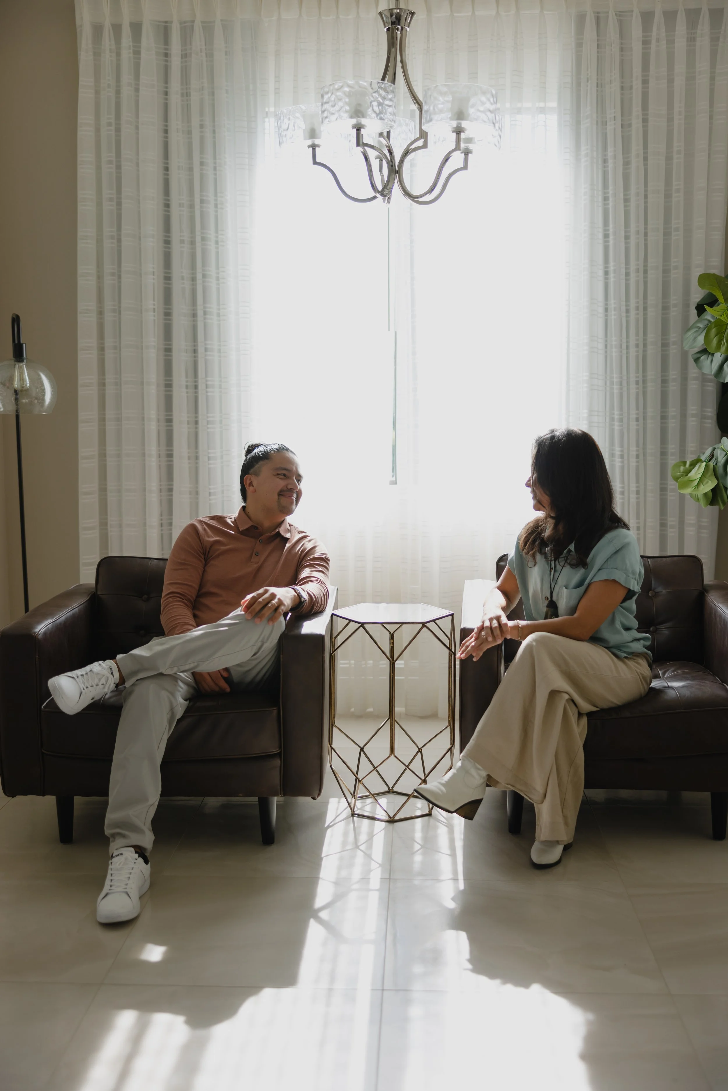 A man and woman sitting on black leather sofas facing each other, chatting in a bright room with white curtains and a chandelier overhead.