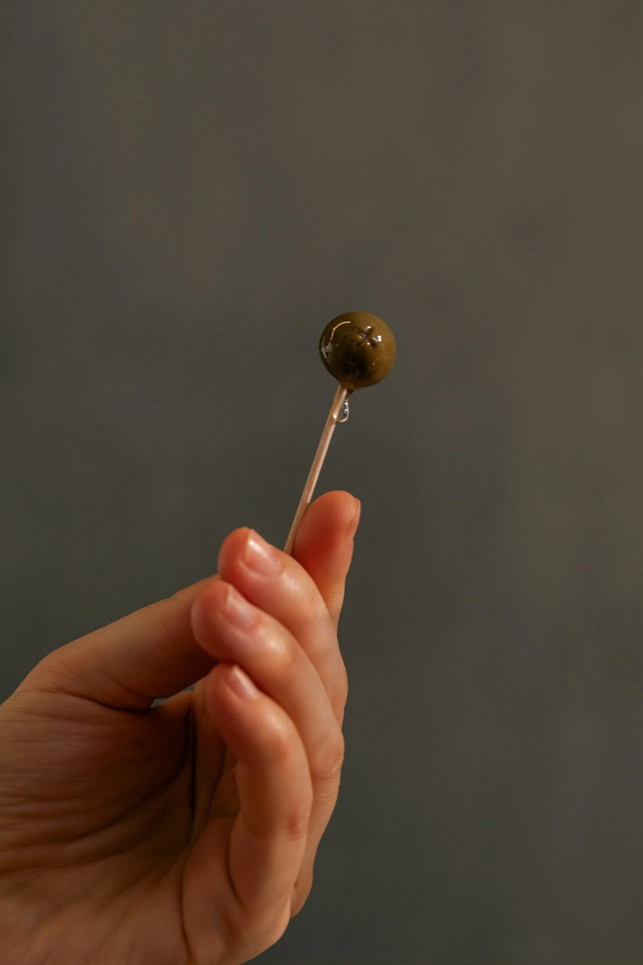 Hand holding a toothpick with a green olive on top against a gray background.