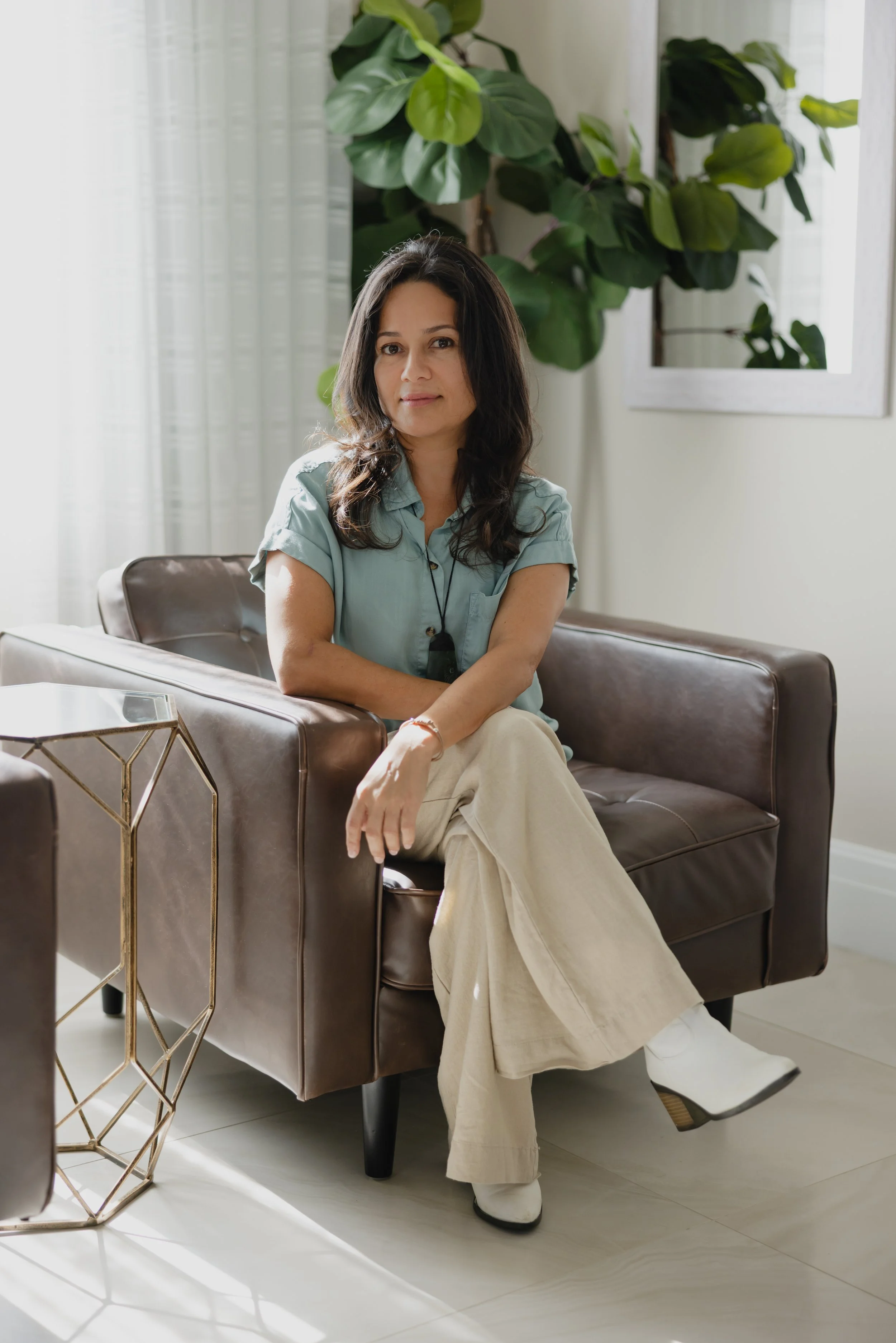 A woman with long dark hair sitting on a brown leather couch in a bright room with large green plants. She is wearing a light blue shirt, beige pants, and white shoes.