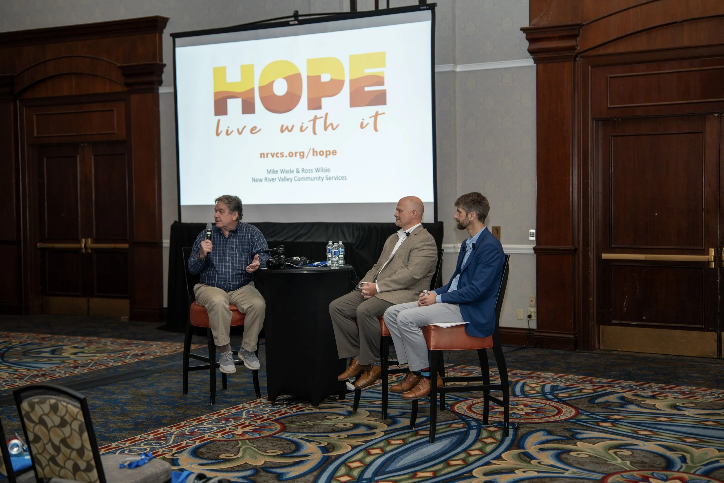 Three men recording a live podcast in front of a screen reading "HOPE. Live with it."