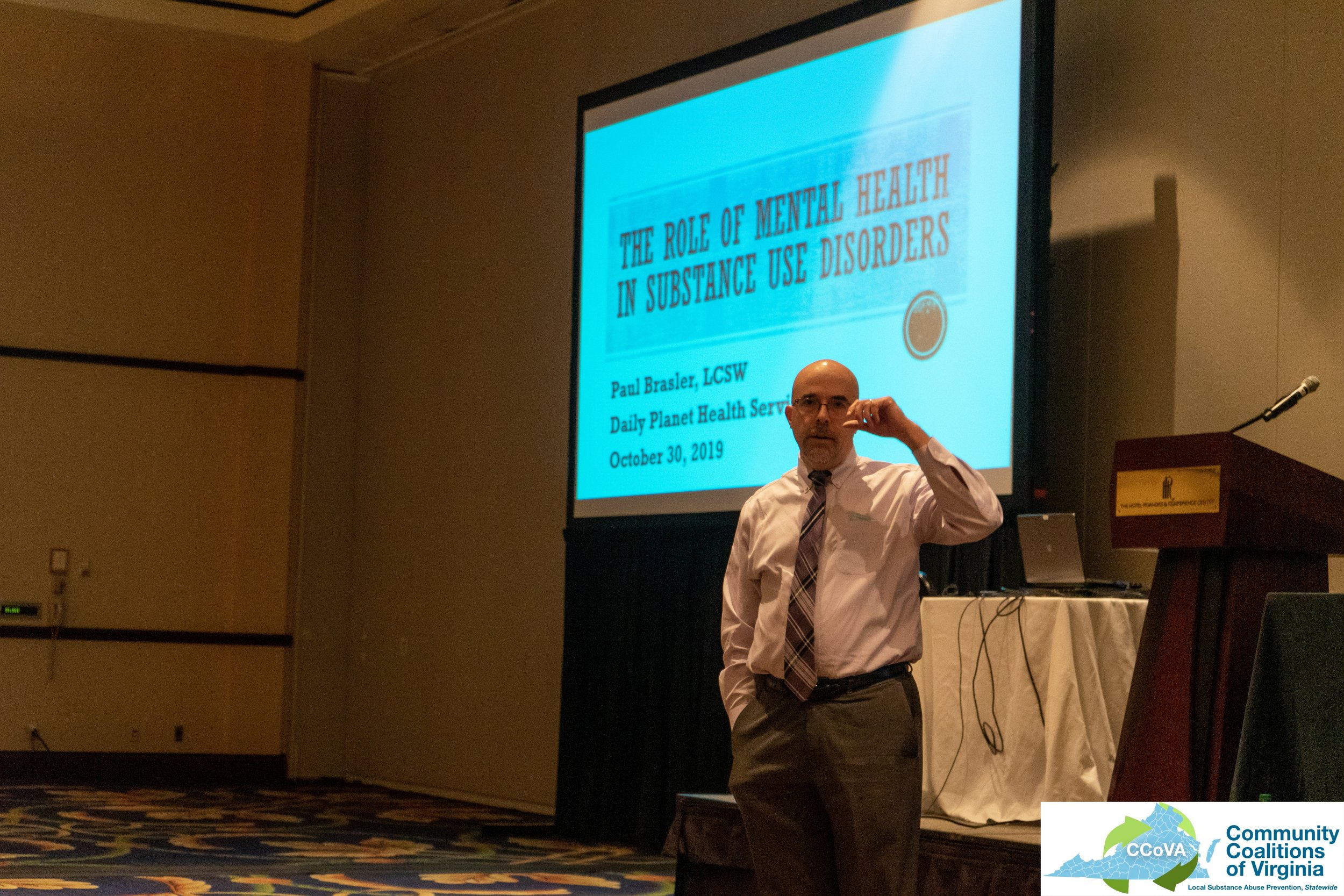 Presenter speaking in front of projector screen with title "The Role of Mental Health in Substance Use Disorders"