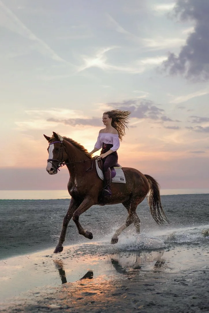 A woman riding a brown horse on a beach at sunset, with the horse splashing through shallow water. The woman is smiling and has long, flowing hair. The sky is partly cloudy with pastel colors. K.N. Linger
