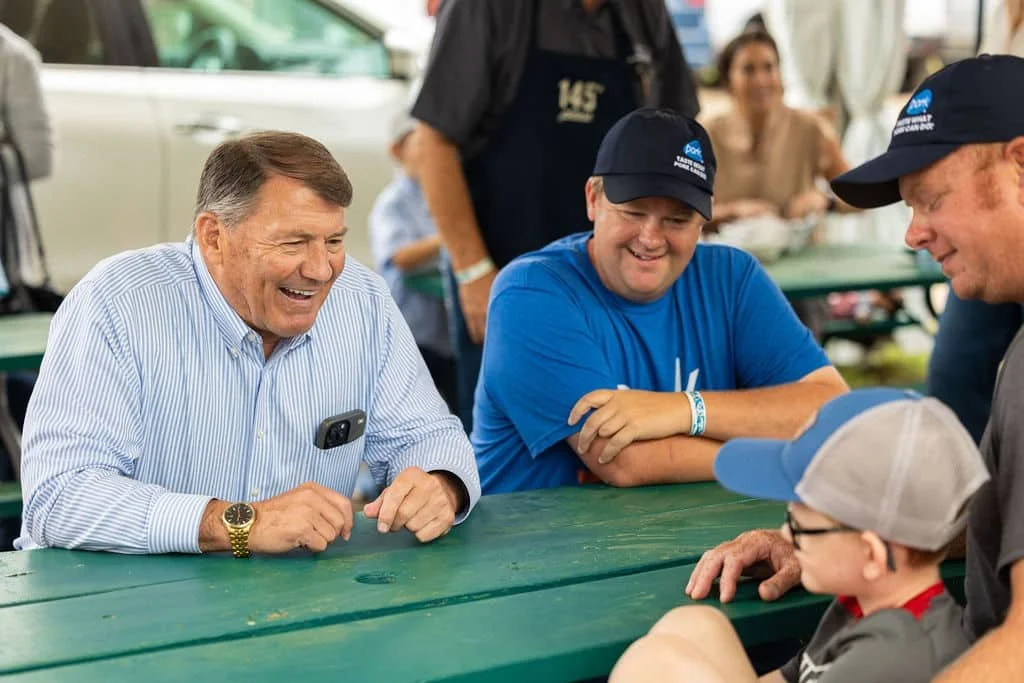 A group of smiling people, including an older man, two boys, and another man, gather around a green picnic table outdoors. They appear to be enjoying a conversation, with the older man and boys engaging warmly.