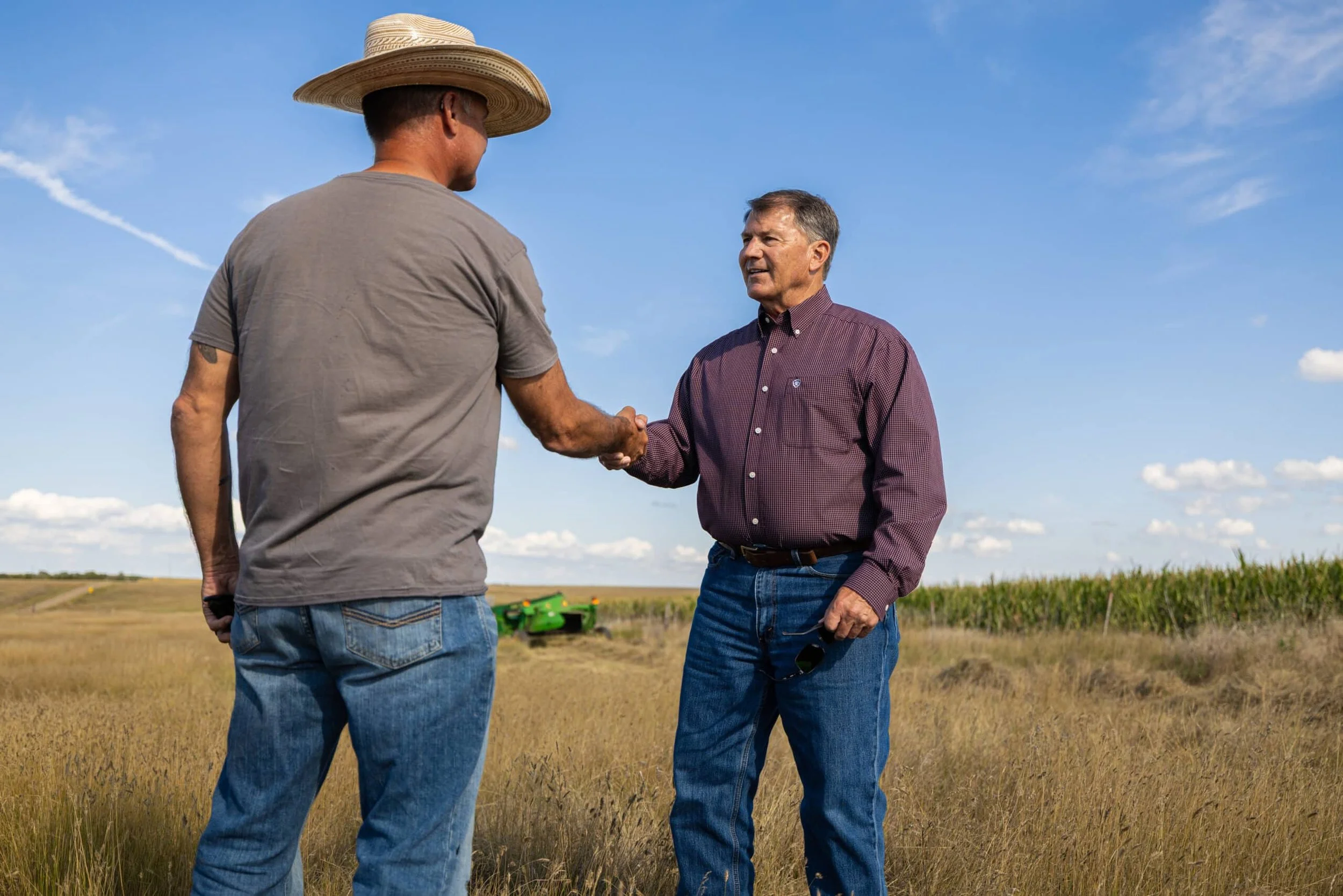 Two men shaking hands in a field under blue sky, one wearing a wide-brimmed hat and casual clothes, the other in a maroon shirt holding sunglasses, with a green tractor in the background.