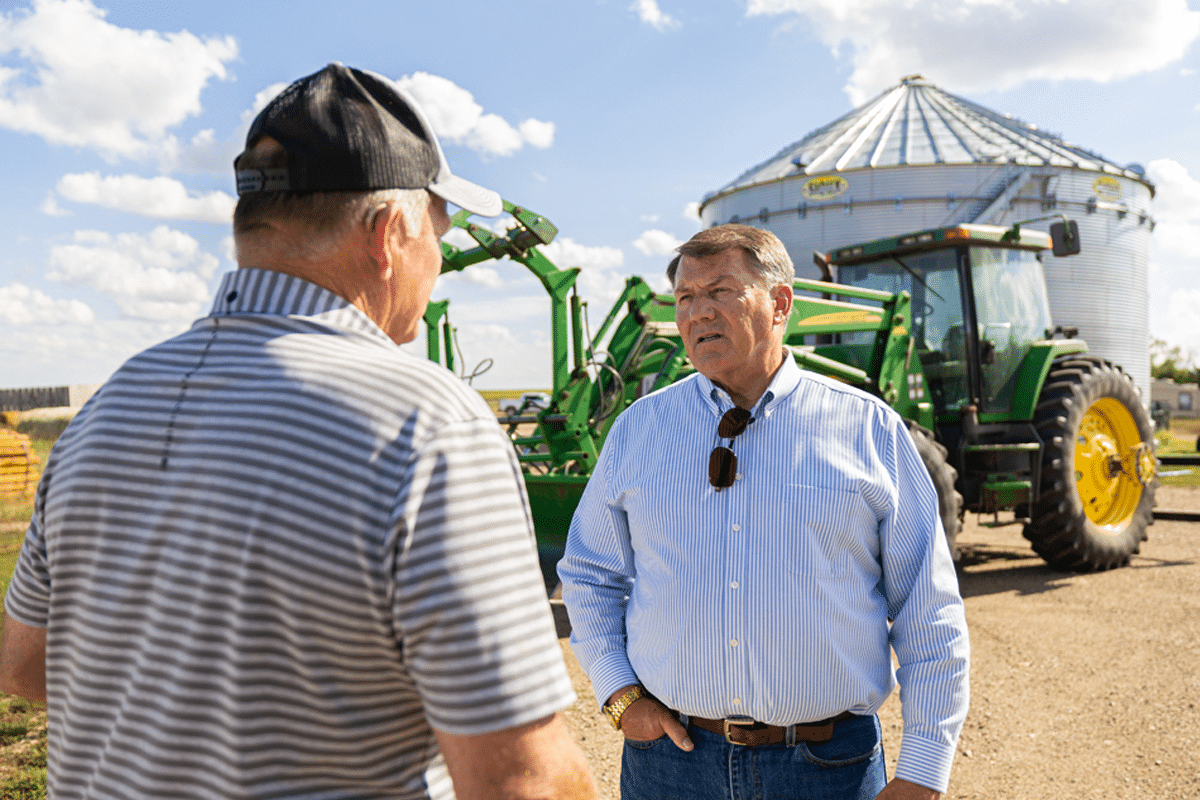 Two men talking outdoors near a large green tractor with a grain silo in the background. One man is wearing a striped shirt and black cap, the other a light blue striped shirt with sunglasses hanging from the collar.