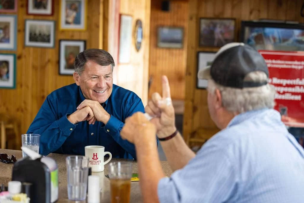 Two men having a conversation in a casual restaurant or cafe, with wood-paneled walls decorated with framed photos. One man is smiling and resting his chin on his hands, while the other, wearing a cap, is gesturing with his finger.