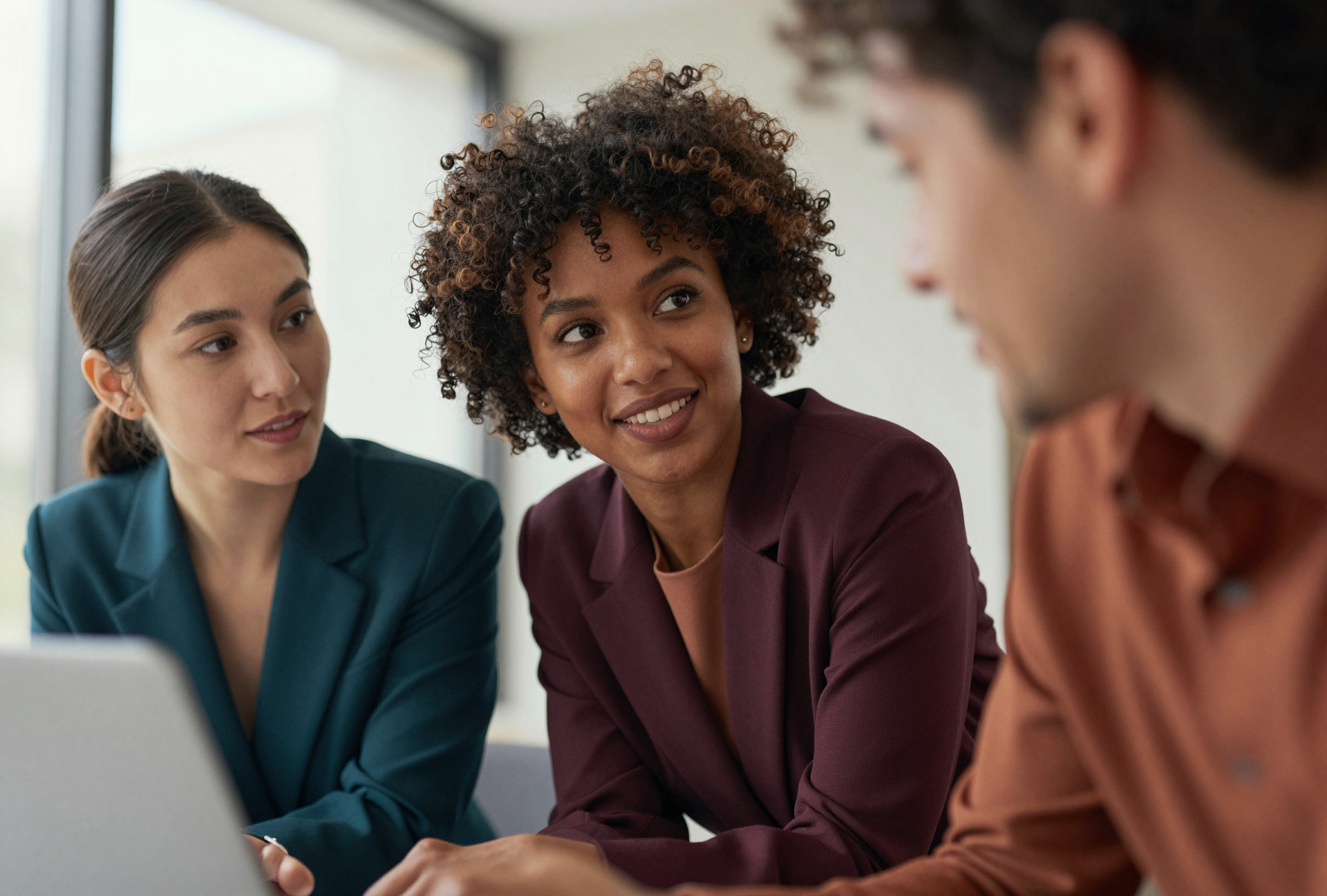 Three women engaged in conversation in a professional setting, sitting near a window, with a laptop on the table.
