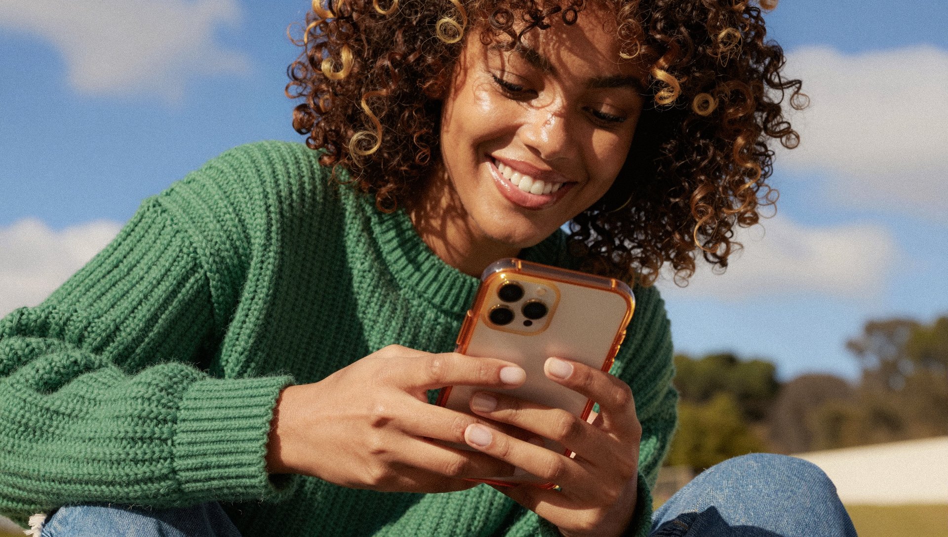 Woman in green sweater sitting outdoors, smiling while looking at her smartphone.